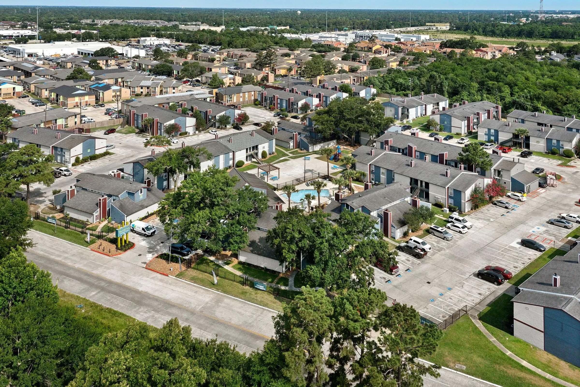 Aerial view of a residential neighborhood featuring several apartment buildings arranged in clusters, with green trees and landscaped areas. A swimming pool is visible in the center of the complex, surrounded by parked cars, pathways, and well-maintained lawns, showcasing a mix of urban living and greenery.