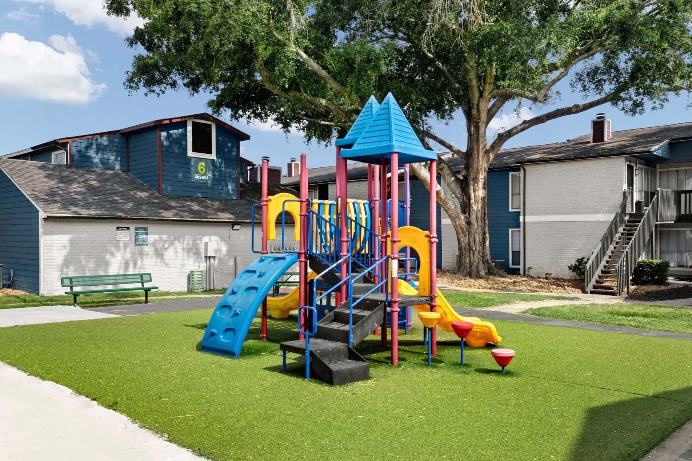 A colorful playground featuring a slide, climbing structure, and various play equipment situated on green artificial grass. Surrounding the playground are residential buildings and a bench nearby, with trees and blue skies in the background.