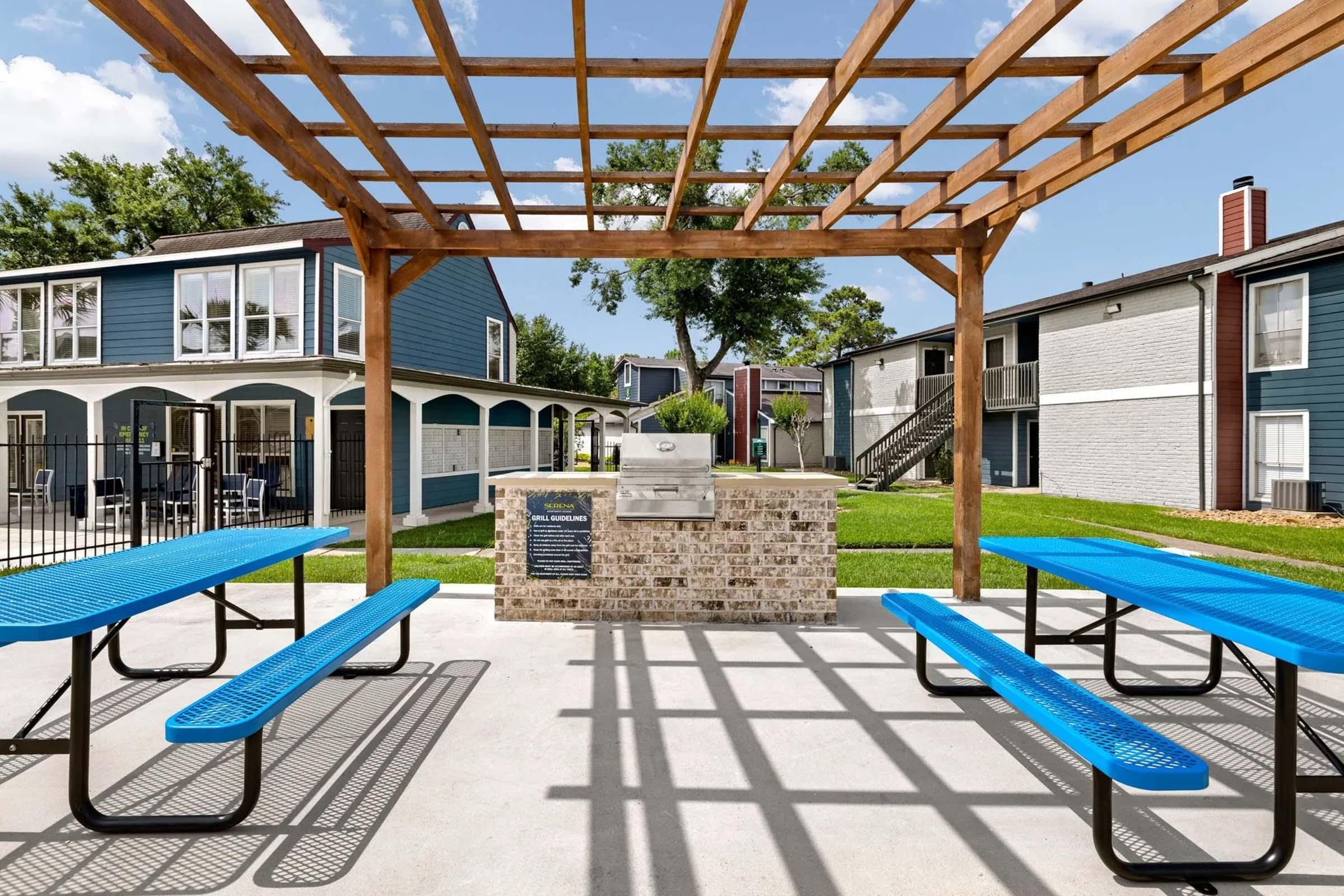 A shaded outdoor grilling area with a stone grill and blue picnic tables under a wooden pergola. The background features a well-maintained lawn and several apartment buildings, with clear skies above. The space is designed for social gatherings and outdoor cooking.
