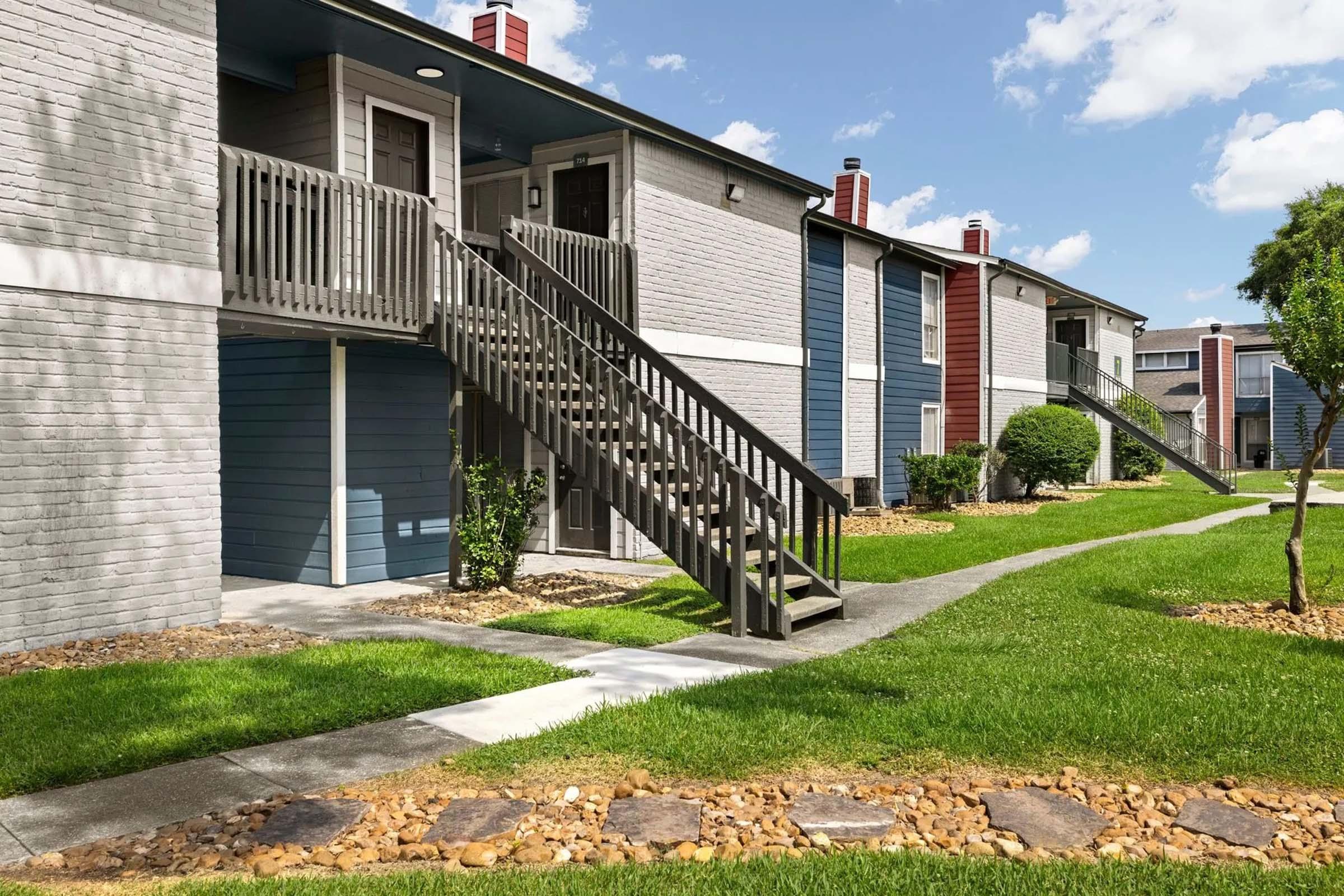 Exterior view of a multi-unit residential complex featuring two-story buildings with balconies, wooden staircases, green lawns, and landscaped pathways. The buildings have a mix of gray and colored siding, and there are trees and shrubs enhancing the outdoor space. Blue sky with scattered clouds is visible.
