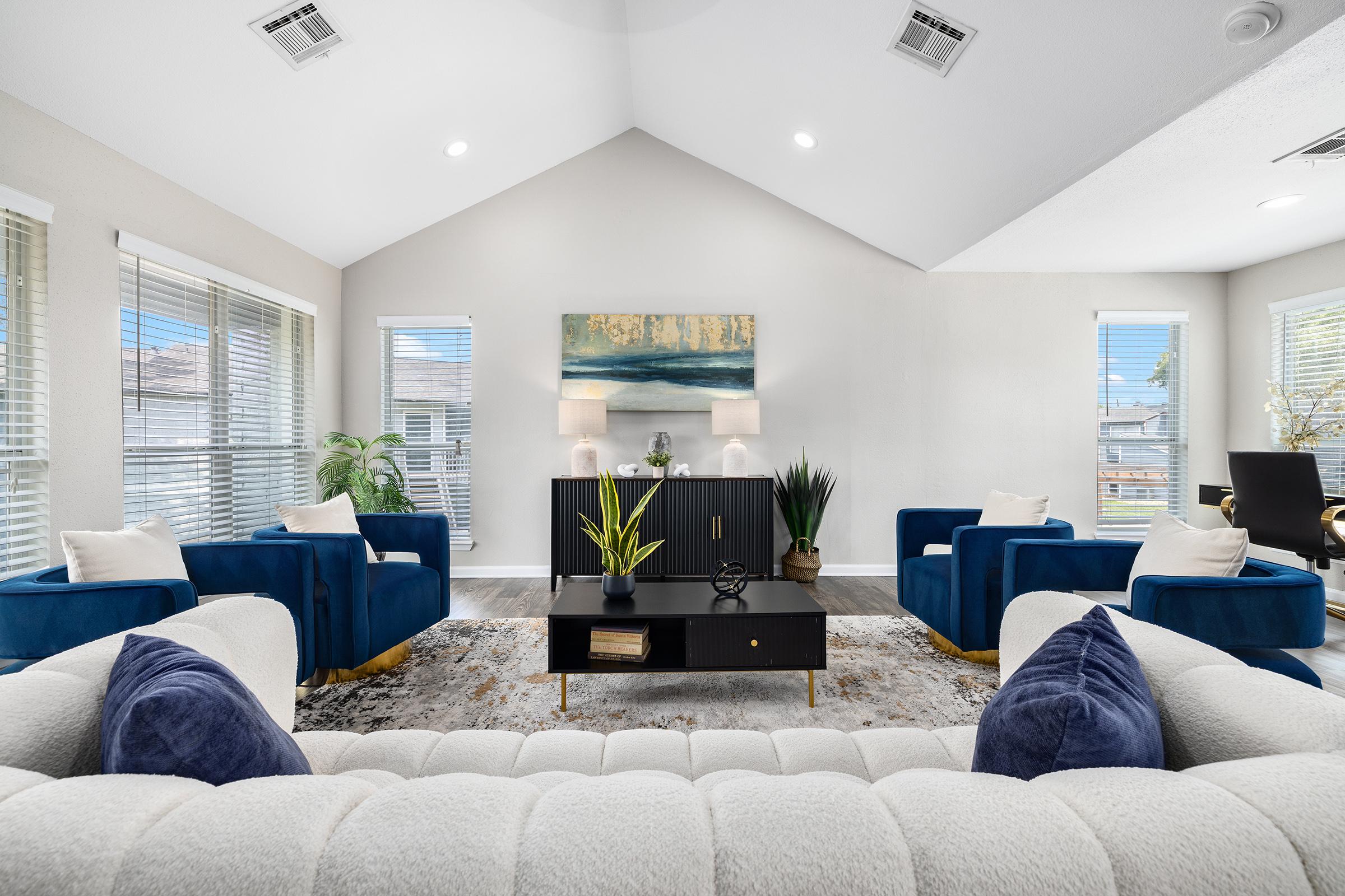 Modern living room featuring a high ceiling, light-colored walls, and large windows allowing natural light. The space includes a cream-colored sofa with decorative pillows, two blue armchairs, a black console table with a plant, and a wall art piece above. A cozy carpet adds warmth to the decor.