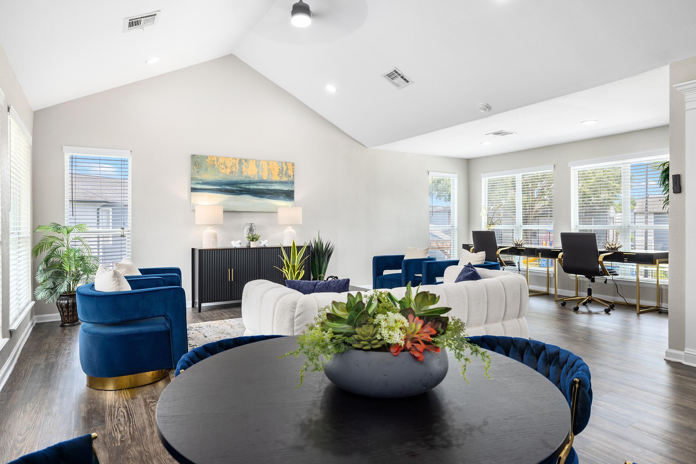 Modern living room featuring a spacious layout with high ceilings. It includes a stylish white sofa, blue accent chairs, a round dining table with a decorative centerpiece, and a sleek sideboard. Large windows allow natural light, and there are potted plants for added greenery. A separate workspace area is visible.