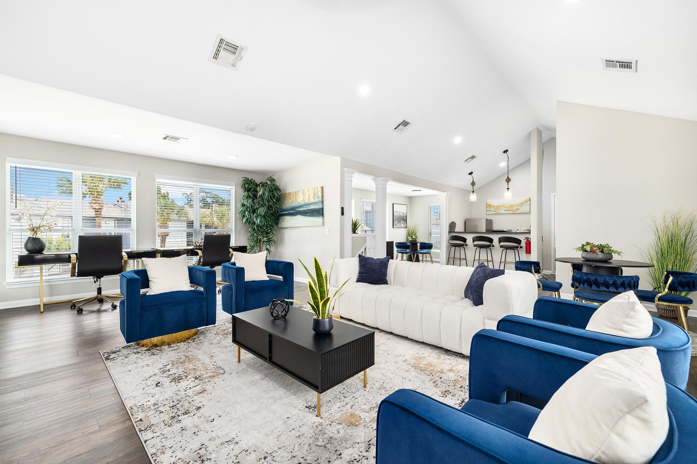 A modern living room with a large white sectional sofa and blue accent chairs. A coffee table sits in the center, surrounded by a light-colored rug. Large windows let in natural light, and there are plants and decor elements throughout. The space is well-lit and open, creating a welcoming atmosphere.