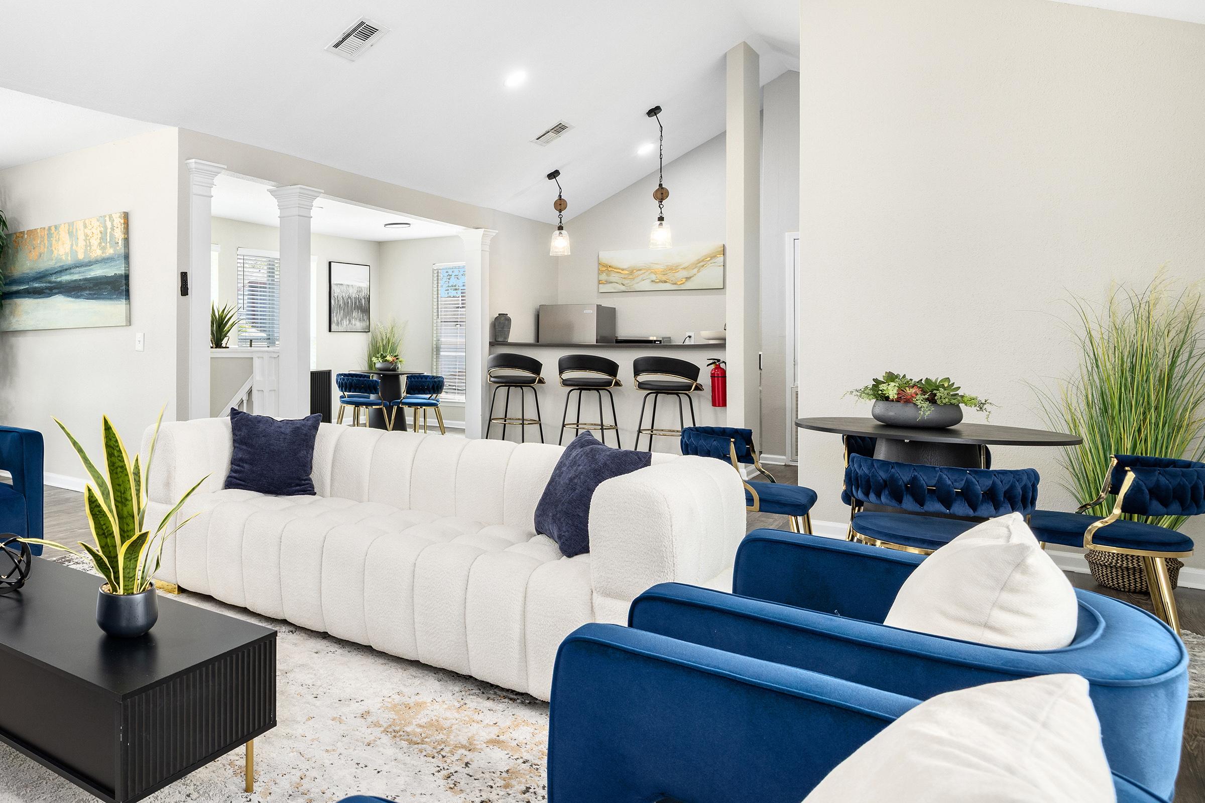 A modern living space featuring a plush white sofa with navy blue accent chairs, a coffee table, and a stylish black console. A dining area with high-backed chairs is visible in the background, along with a kitchen bar. Decorative plants and artwork adorn the space, creating a bright and inviting atmosphere.