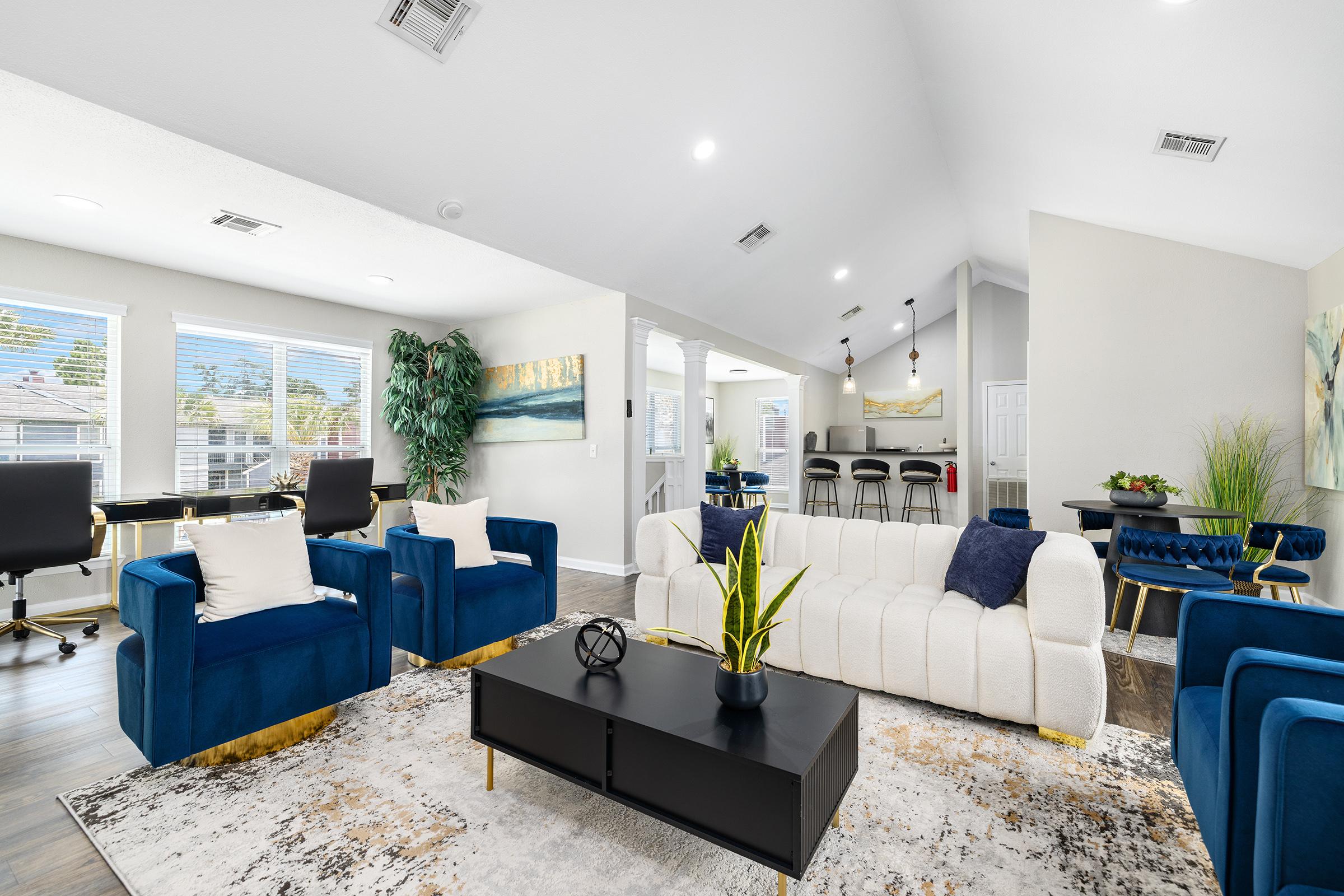 A modern living room featuring blue armchairs and a white sofa with decorative pillows. A black coffee table is in the center, surrounded by a large area rug. There are plants and wall art, and the space is well-lit with large windows. The room leads to a dining area with a light and airy feel.