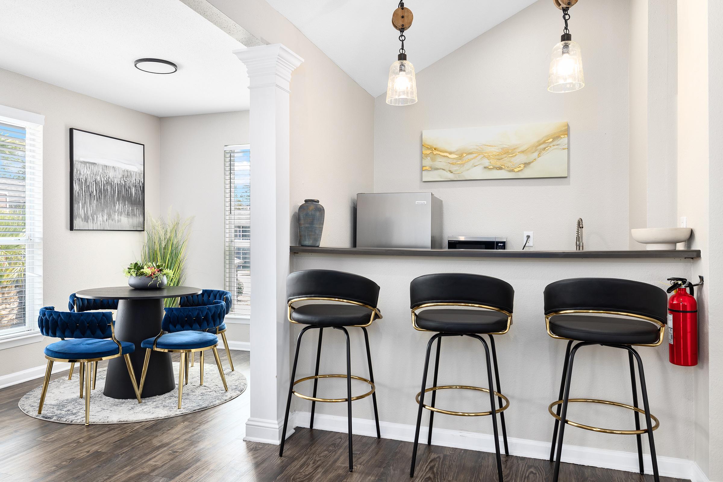 A modern dining area featuring a round black table with blue velvet chairs, surrounded by large windows. Nearby, a sleek grey kitchen bar with three black and gold stools, a small fridge, and decorative artwork above the bar. A fire extinguisher is mounted on the wall.