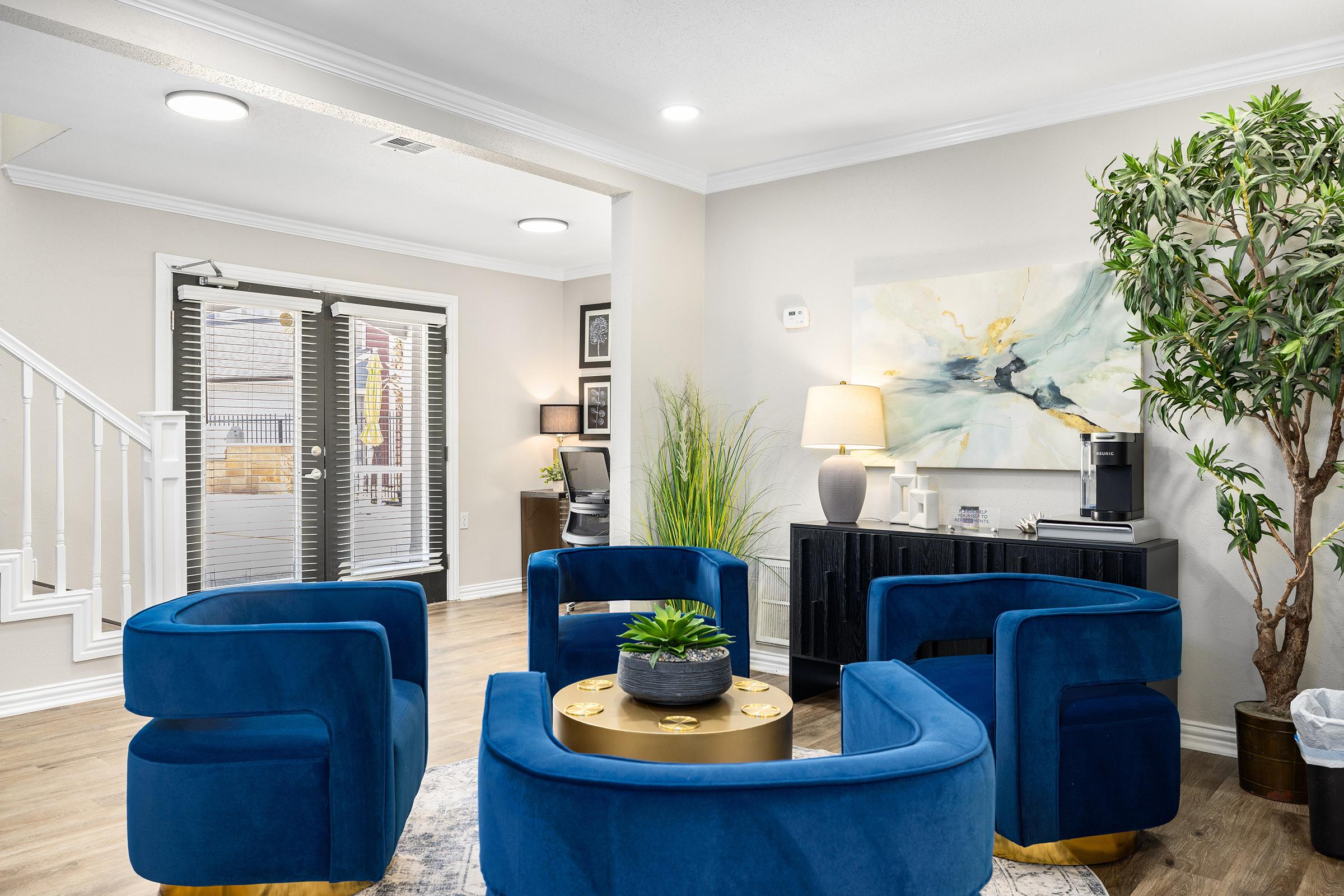 A cozy living area featuring modern blue velvet chairs arranged around a circular gold coffee table, with a green plant centerpiece. The walls are painted in a light color, and there's a large abstract painting on one wall. A lamp and a dark cabinet with a coffee maker add to the inviting atmosphere.
