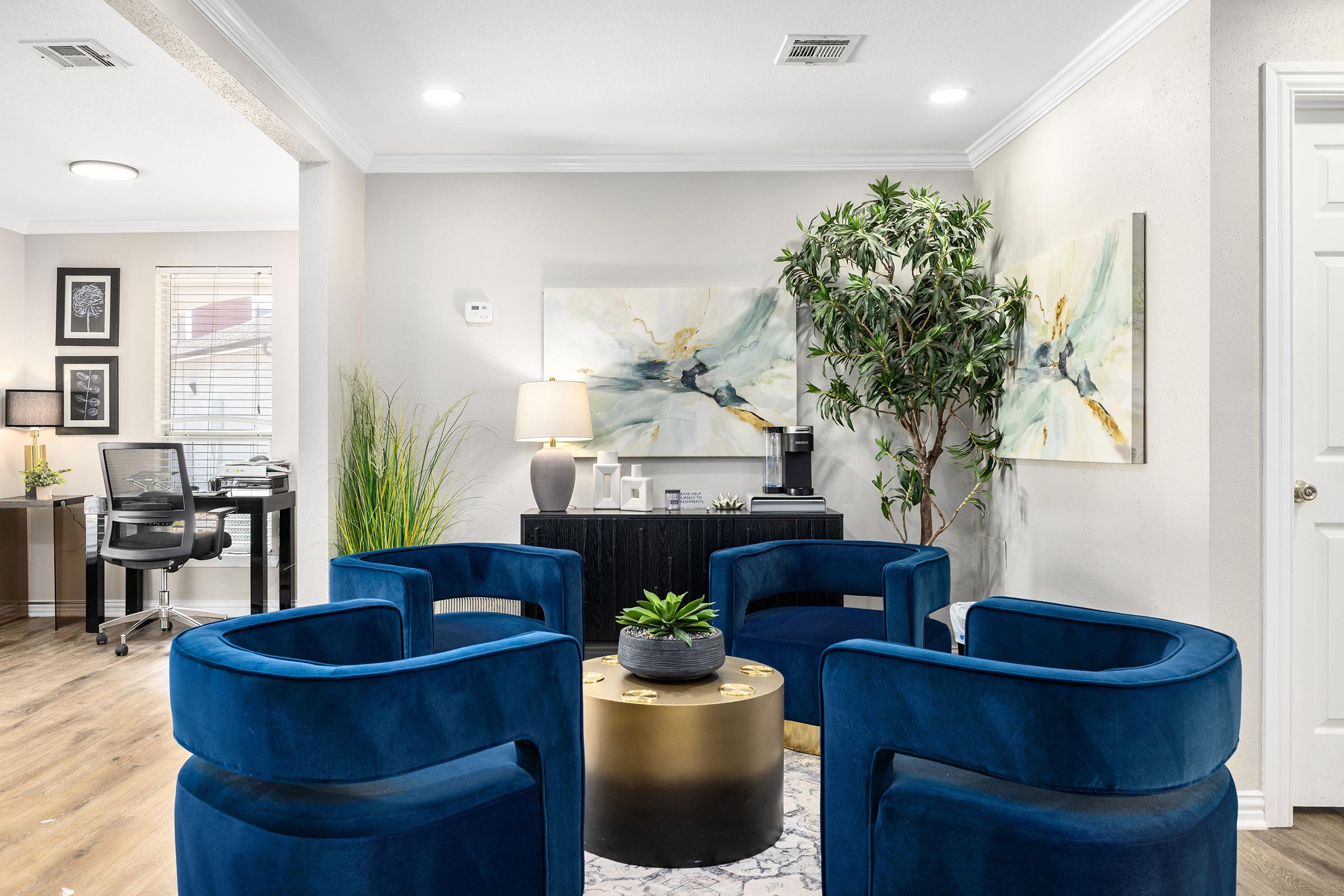 A modern living space featuring four blue velvet chairs arranged around a gold and white coffee table. In the background, there are two abstract paintings on the walls, a potted plant, and a small coffee station. A home office is visible through an open door, adding a functional element to the cozy atmosphere.