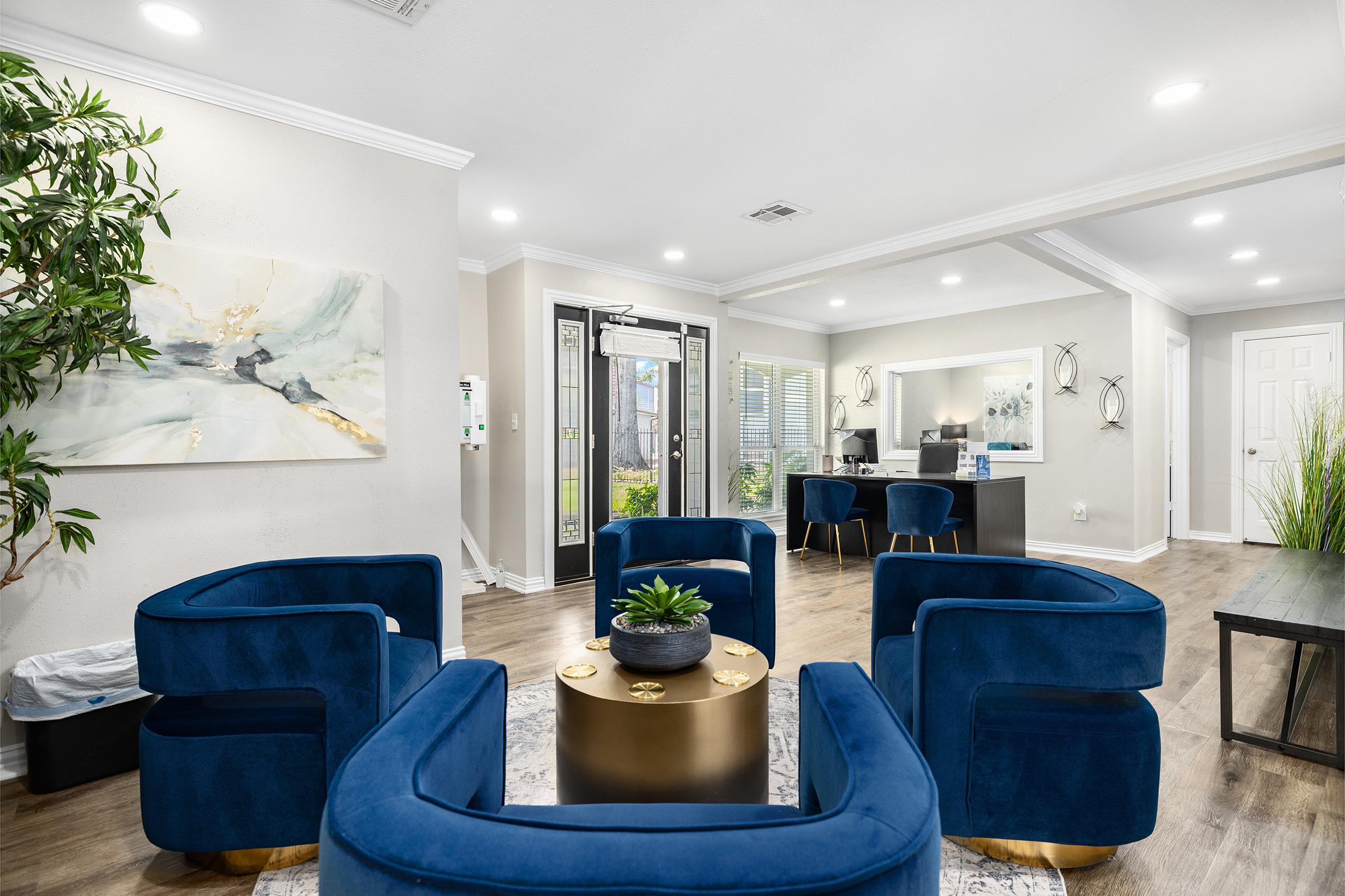 A modern living space featuring blue upholstered armchairs arranged around a round gold coffee table. A potted plant sits on the table. The room has a painting on the wall, large windows with light filtering in, and a desk area with chairs visible in the background.