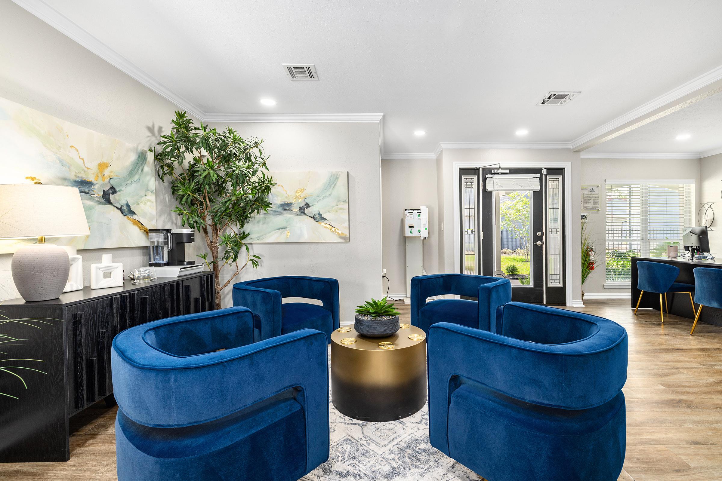 A modern waiting area featuring blue upholstered chairs arranged around a round coffee table with a small plant. The walls are adorned with abstract artwork, and there's a console table with decorative items. Large windows allow natural light, and visible greenery is outside. An inviting and stylish atmosphere.