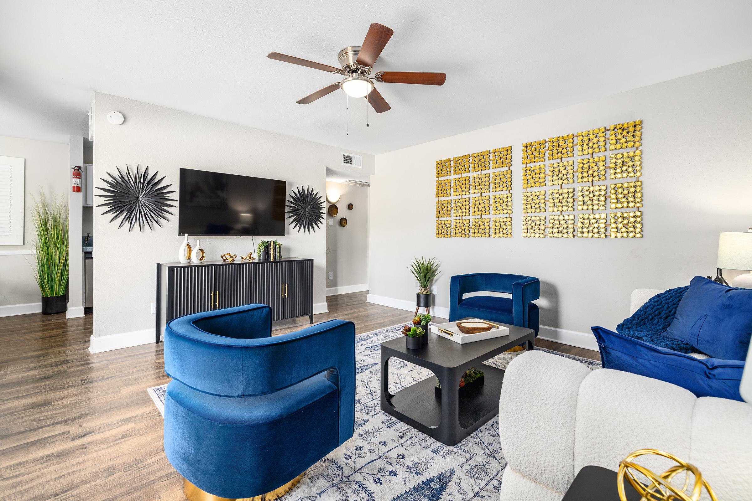 A stylish living room featuring a ceiling fan, a large flat-screen TV on a dark media console, and two modern blue chairs. A decorative wall with gold geometric art complements the space. The room has hardwood flooring and a cozy atmosphere with plants and decorative accents.