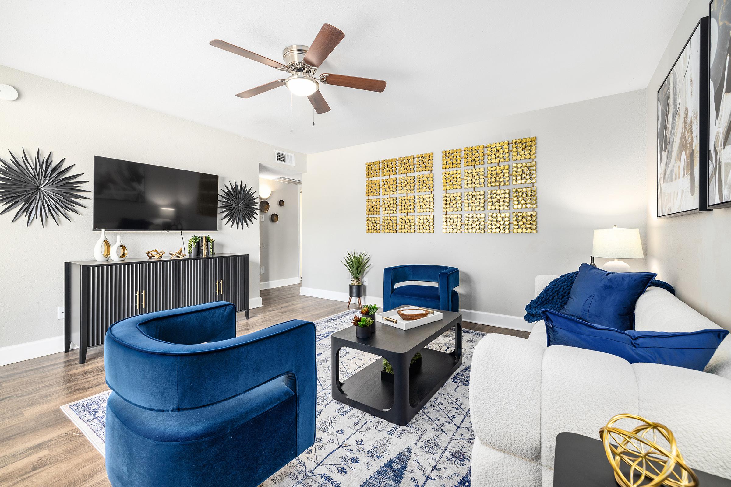A modern living room featuring a stylish light-colored sofa, two blue accent chairs, and a black entertainment unit against a wall with a large flat-screen TV. Decorative wall art and a gold geometric display add elegance. A coffee table is centered on a patterned rug, with plants for a touch of greenery.