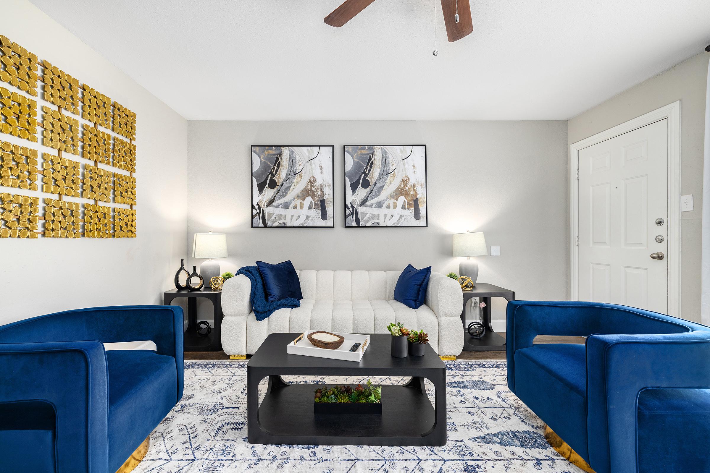 A modern living room featuring a white sofa with blue and gold accent pillows, two blue armchairs, a black coffee table with a small plant, and abstract wall art. The decor includes a textured wall piece, a ceiling fan, and a neutral wall color, creating a cozy and stylish atmosphere.