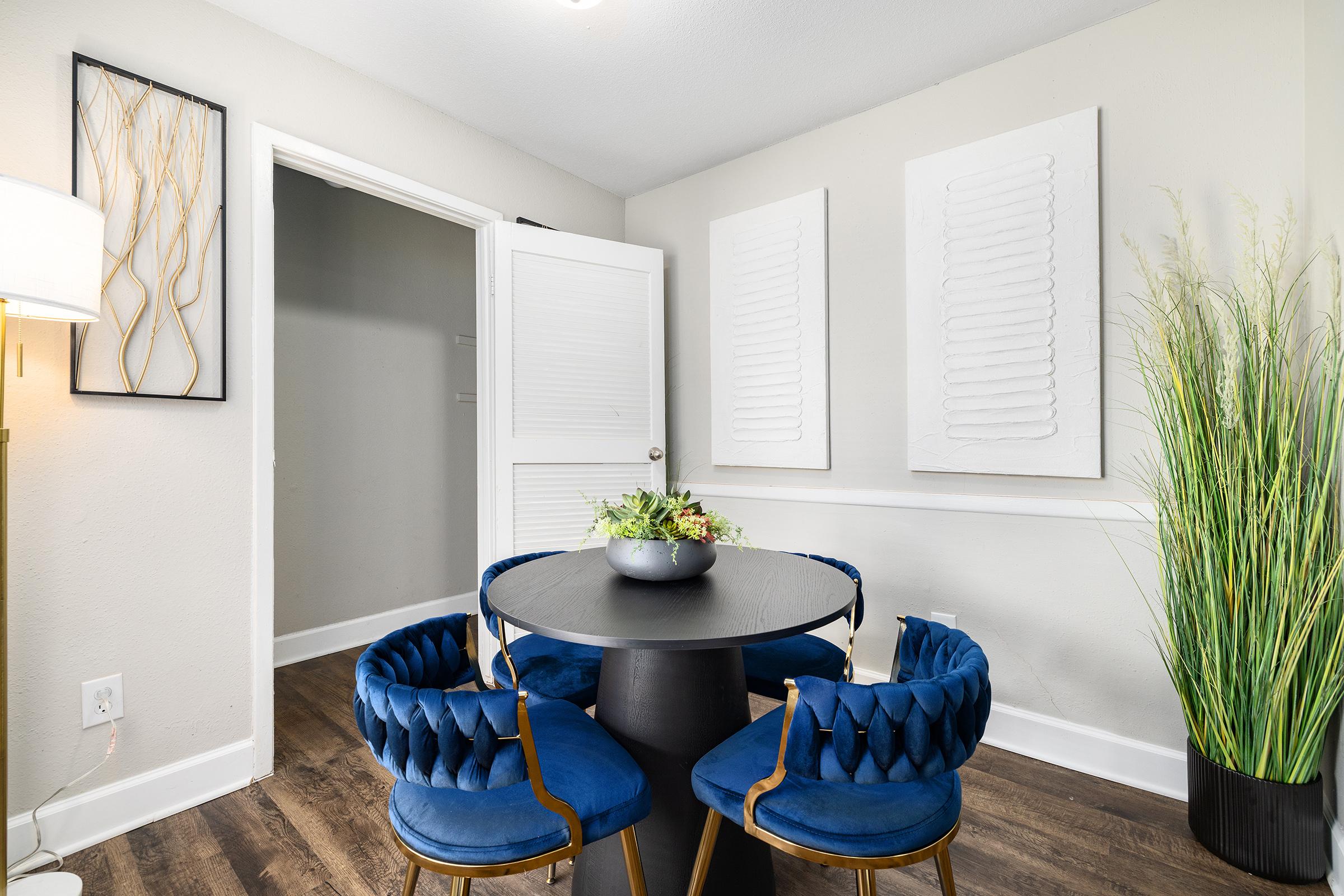 A cozy dining area featuring a round black table surrounded by plush blue chairs. The walls are adorned with textured white art pieces. There's a decorative plant in the corner and a light fixture illuminating the space, creating a modern and inviting atmosphere.