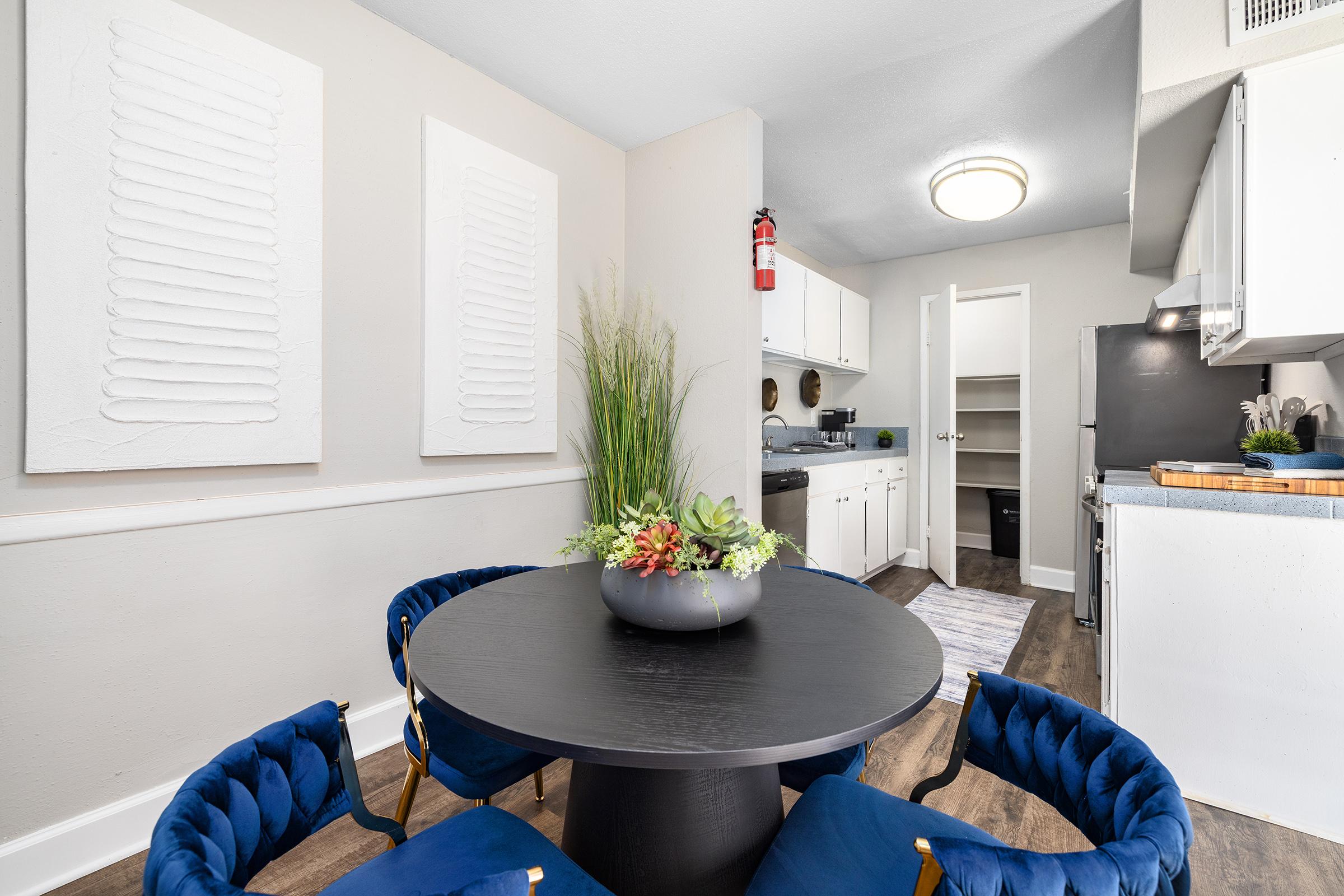 A modern dining area featuring a round black table surrounded by blue velvet chairs. A decorative vase with greenery sits on the table. The light-colored walls have two textured wall art pieces, and a kitchen area is visible in the background with white cabinets and appliances.