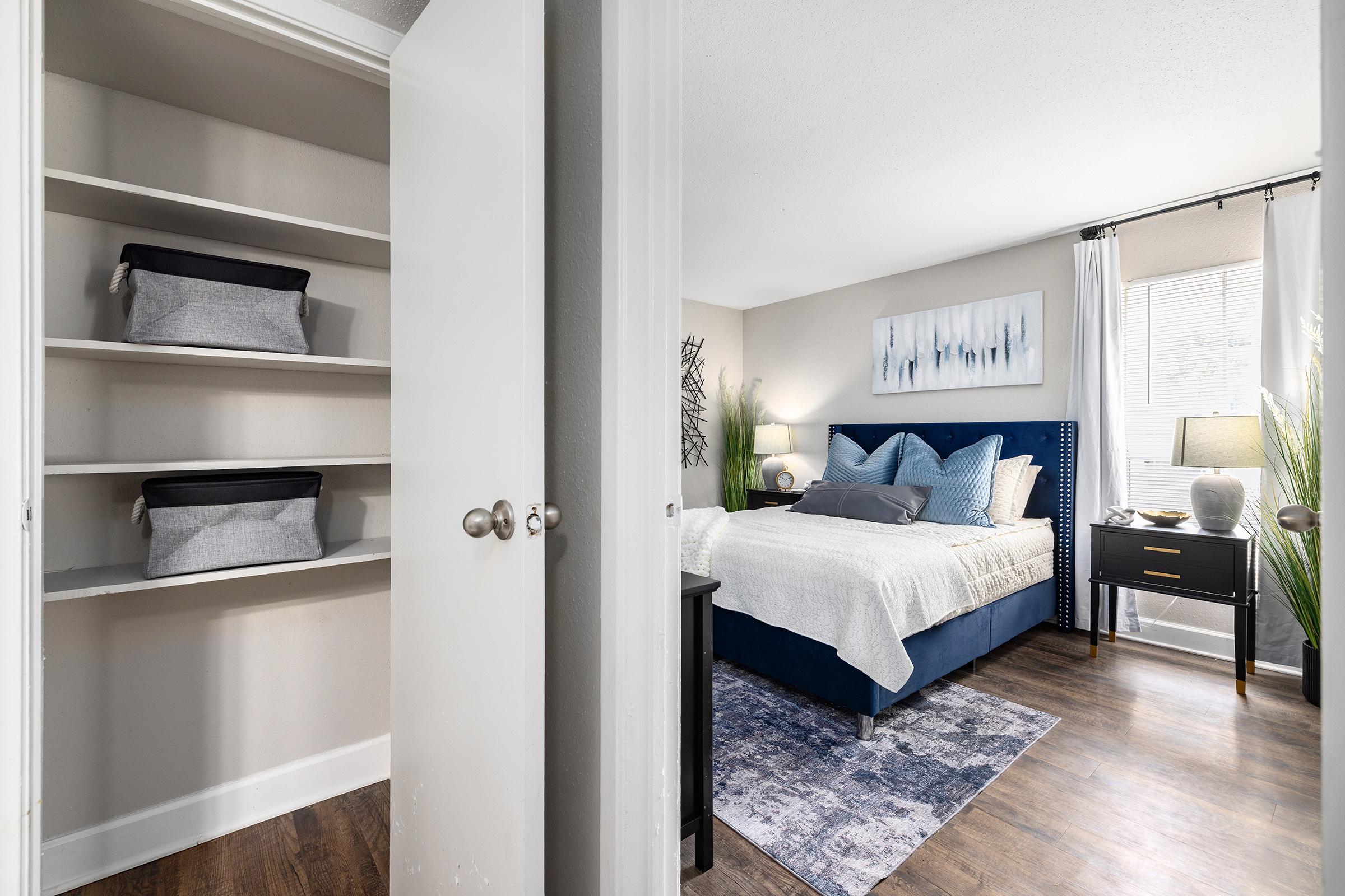 A well-lit bedroom with a blue upholstered bed adorned with decorative pillows, a bedside table with a lamp, and a window with sheer curtains. A closet with neatly organized fabric storage bins is visible on the left side, and a decorative plant adds a touch of greenery to the room.