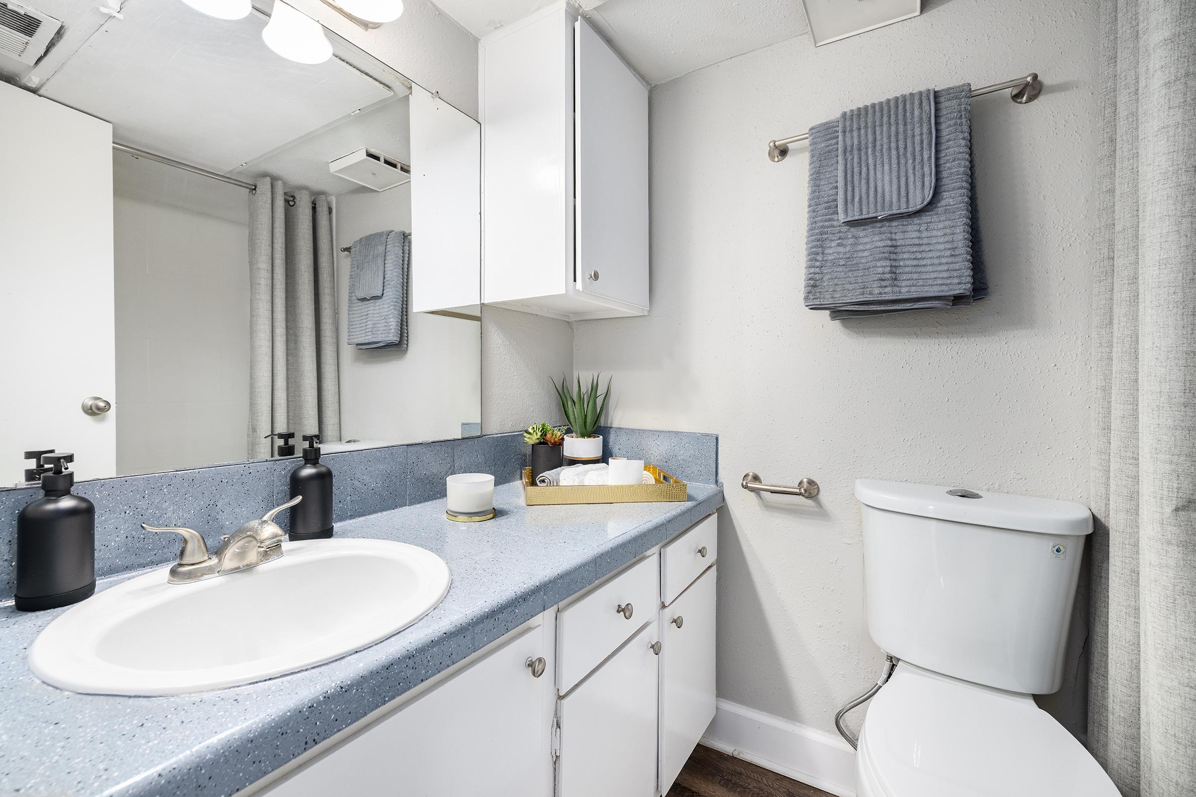 A modern bathroom featuring a white sink with a sleek faucet, a toilet, and a mirror above the sink. The countertop is blue, decorated with a small plant and a tray holding towels and a candle. Grey towels hang on a rack, and there are light-colored curtains partially covering a window.
