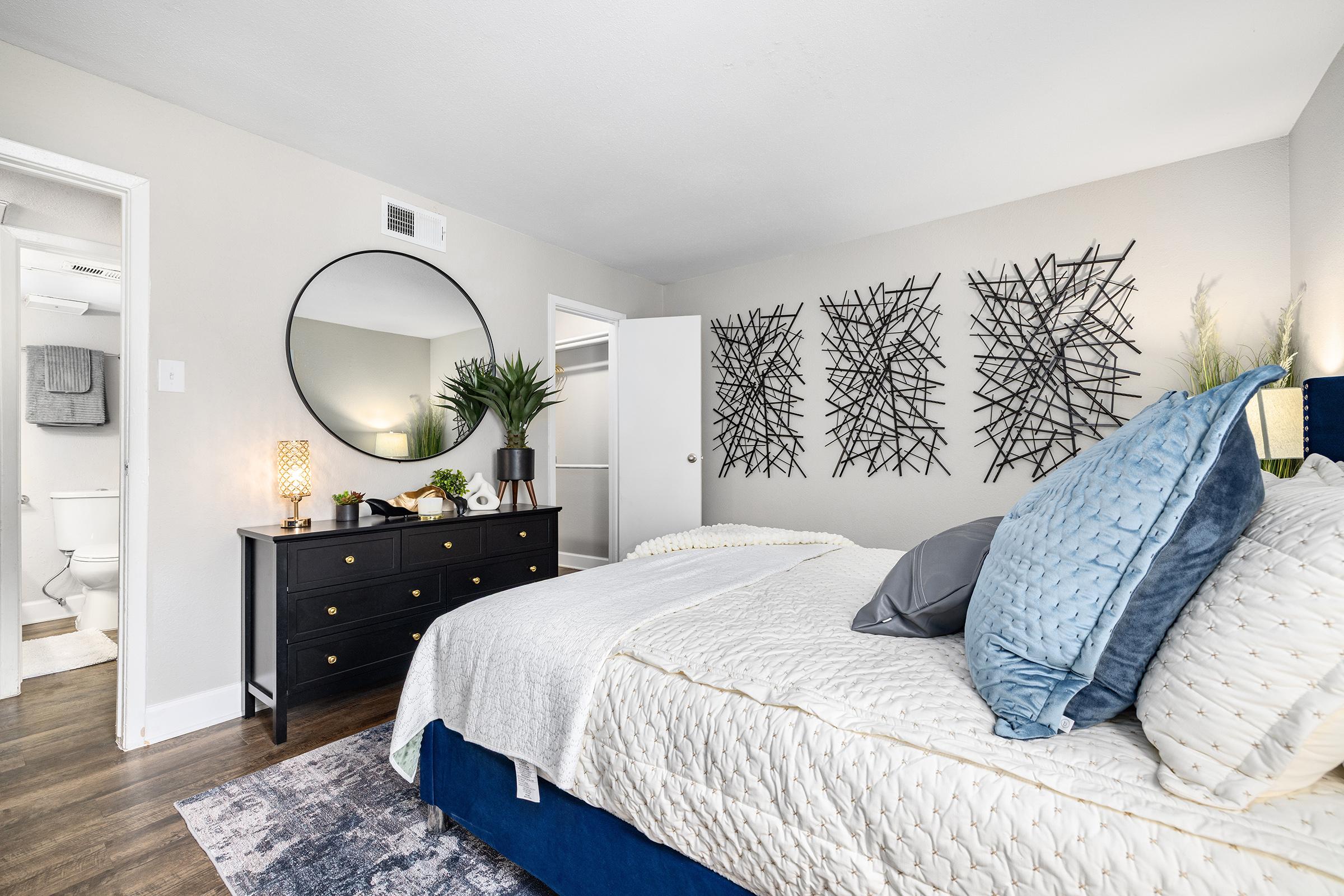 A stylish bedroom featuring a comfortable bed with decorative pillows, a large round mirror above a black dresser, and modern wall art. Natural light illuminates the space, with a potted plant and decorative items on the dresser. A bathroom is visible through an open door.
