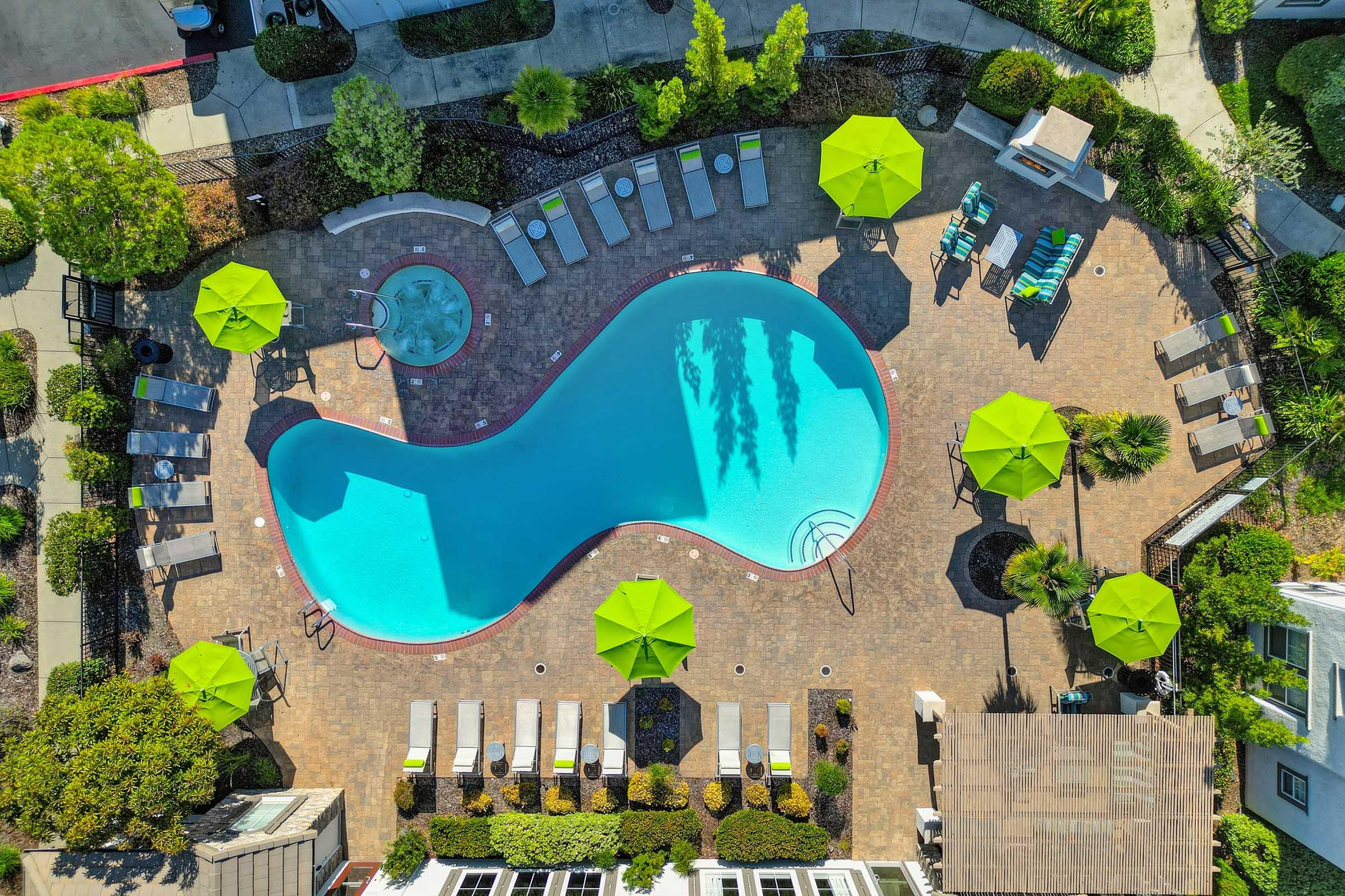 Aerial view of a swimming pool shaped like a figure eight, surrounded by lounge chairs and green umbrellas. Lush landscaping frames the pool area, with a hot tub nearby, and pathways leading to different sections of the property. Bright sunlight illuminates the scene, creating a vibrant, inviting atmosphere.