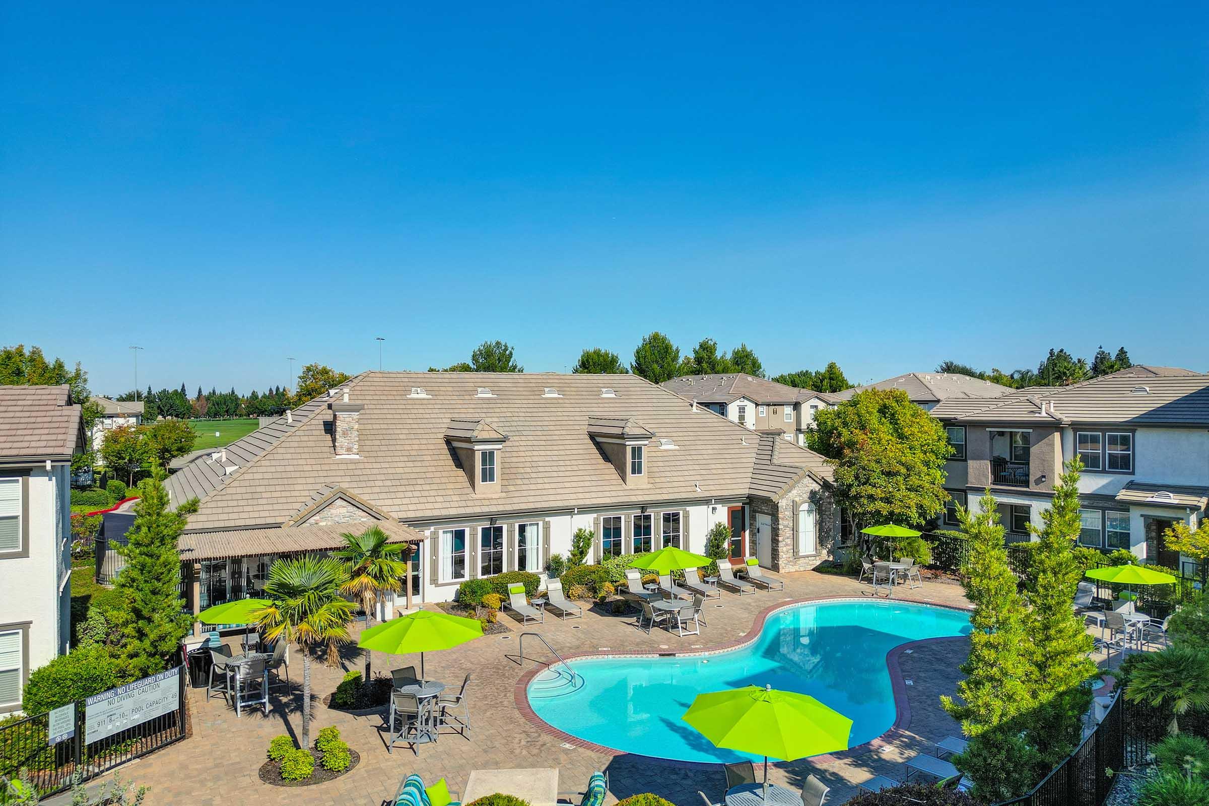 Aerial view of a residential complex featuring a large swimming pool surrounded by green lounge chairs and umbrellas. There are pathways, landscaped areas with trees, and a clubhouse with outdoor seating areas, all under a clear blue sky.