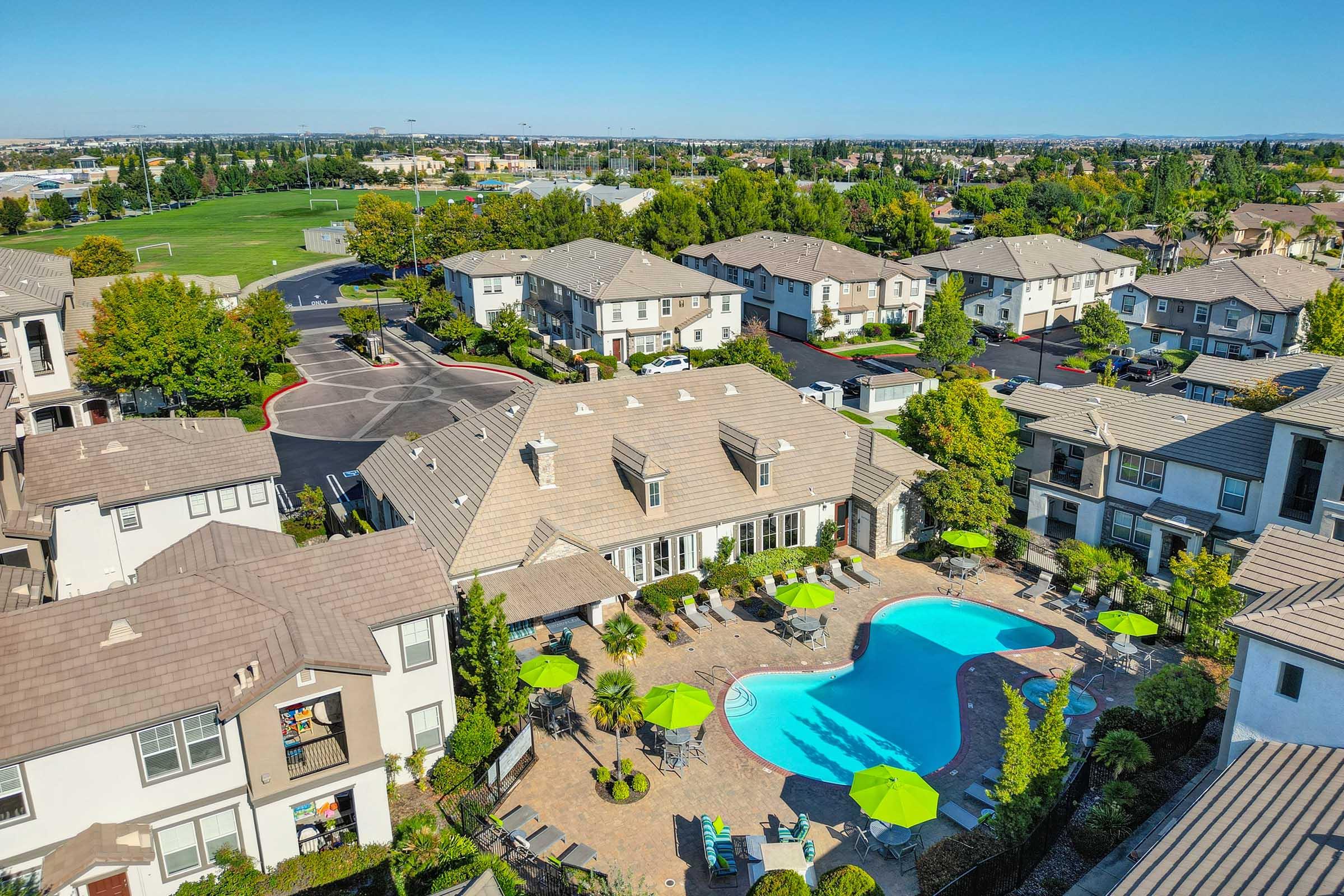 Aerial view of a residential complex featuring multiple buildings, a blue swimming pool surrounded by lounge chairs and umbrellas, and landscaped grounds with trees. In the background, green fields and additional housing can be seen under a clear blue sky.