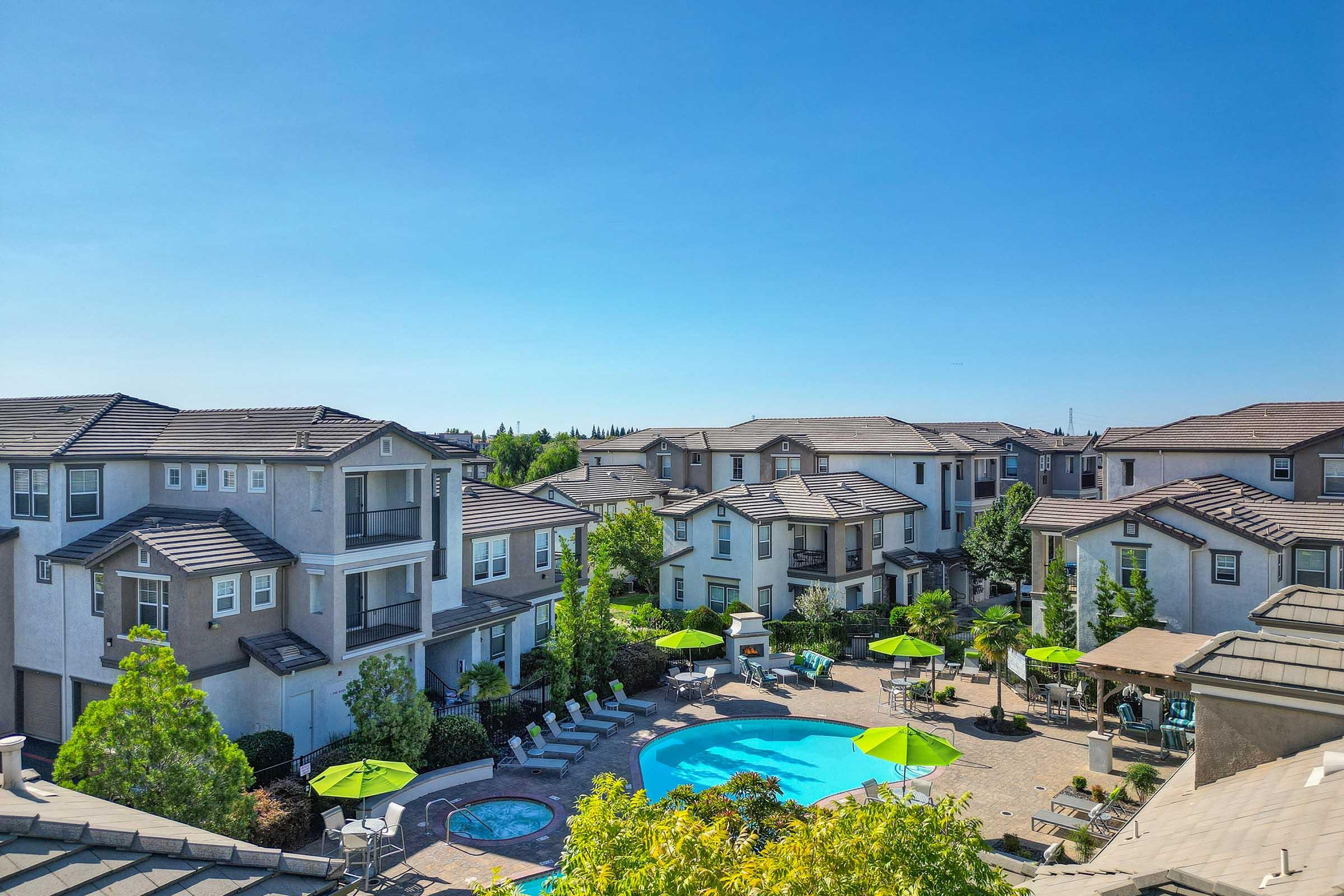 Aerial view of a residential complex featuring multiple buildings surrounding a large swimming pool. The pool area is adorned with green umbrellas and lounge chairs, with landscaped trees and gardens creating a vibrant atmosphere under a clear blue sky.