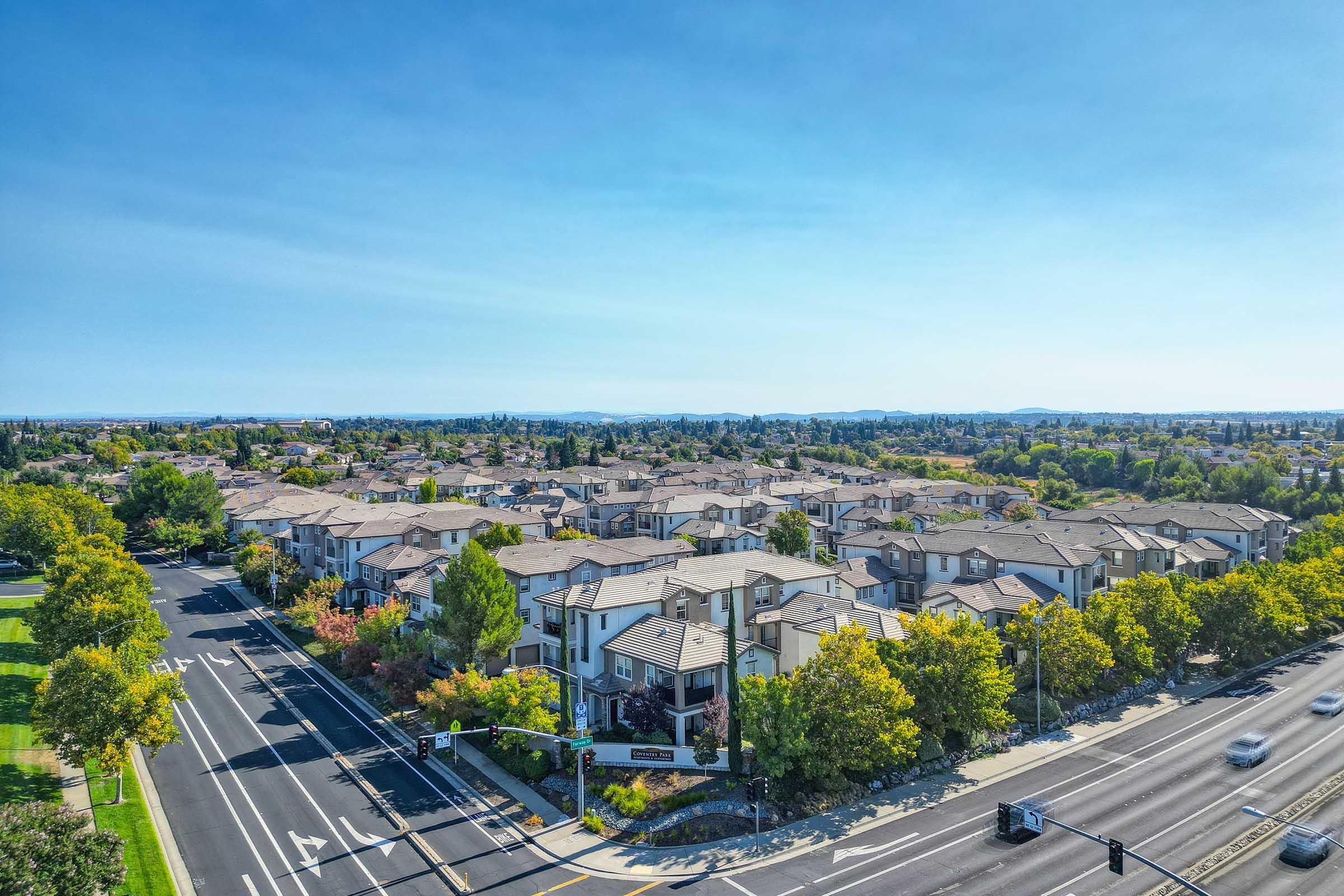 Aerial view of a suburban neighborhood featuring numerous apartment buildings and houses. The area is surrounded by trees and greenery, with clear blue skies overhead. A road runs through the foreground, with traffic lights visible, indicating a well-planned residential community.