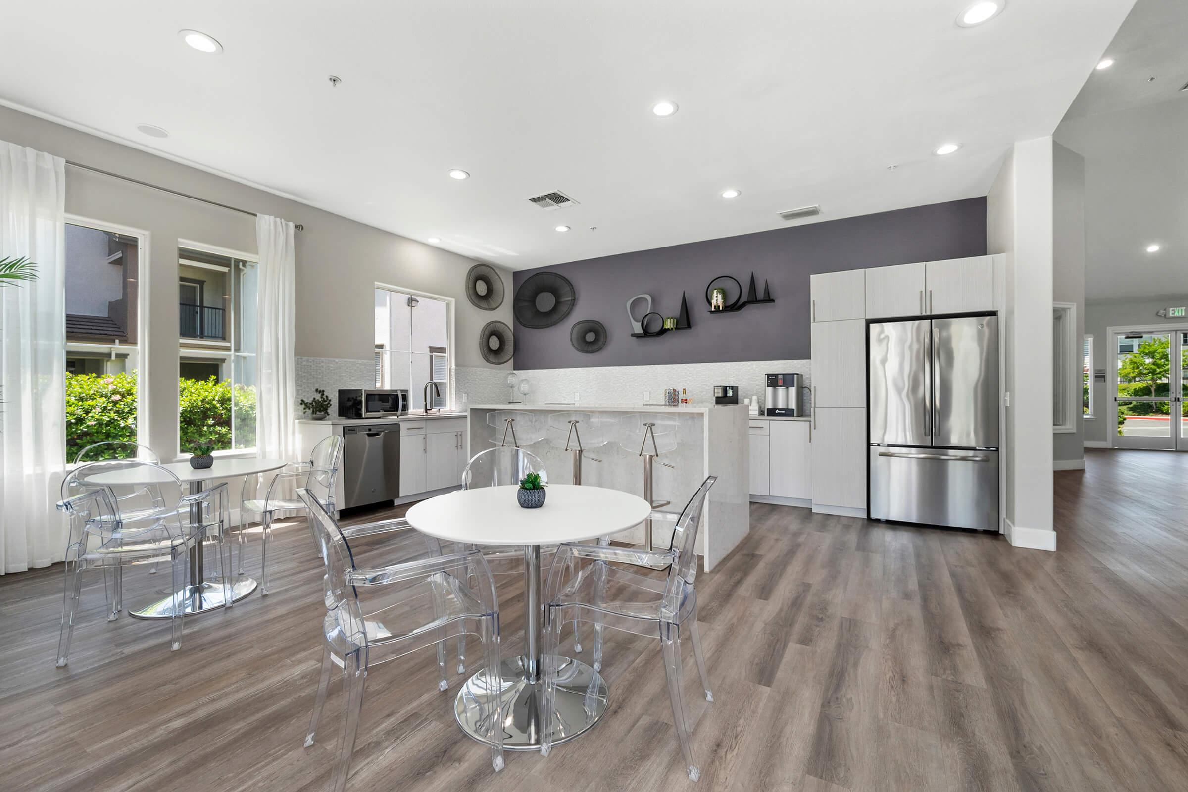 Modern kitchen and dining area featuring a sleek design with a round table and transparent chairs. A countertop with a microwave and coffee station is against the wall, complemented by a stainless steel refrigerator. Bright natural light floods the space through large windows, enhancing the contemporary atmosphere.