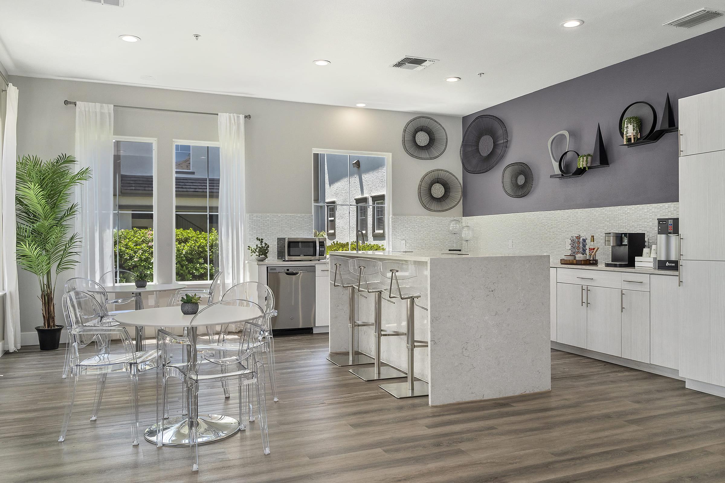 Bright and modern kitchen area featuring a round dining table with clear chairs, bar stools at a sleek countertop, and a variety of appliances including a microwave and coffee maker. Large windows allow natural light, and decorative wall accents add a contemporary touch to the space.