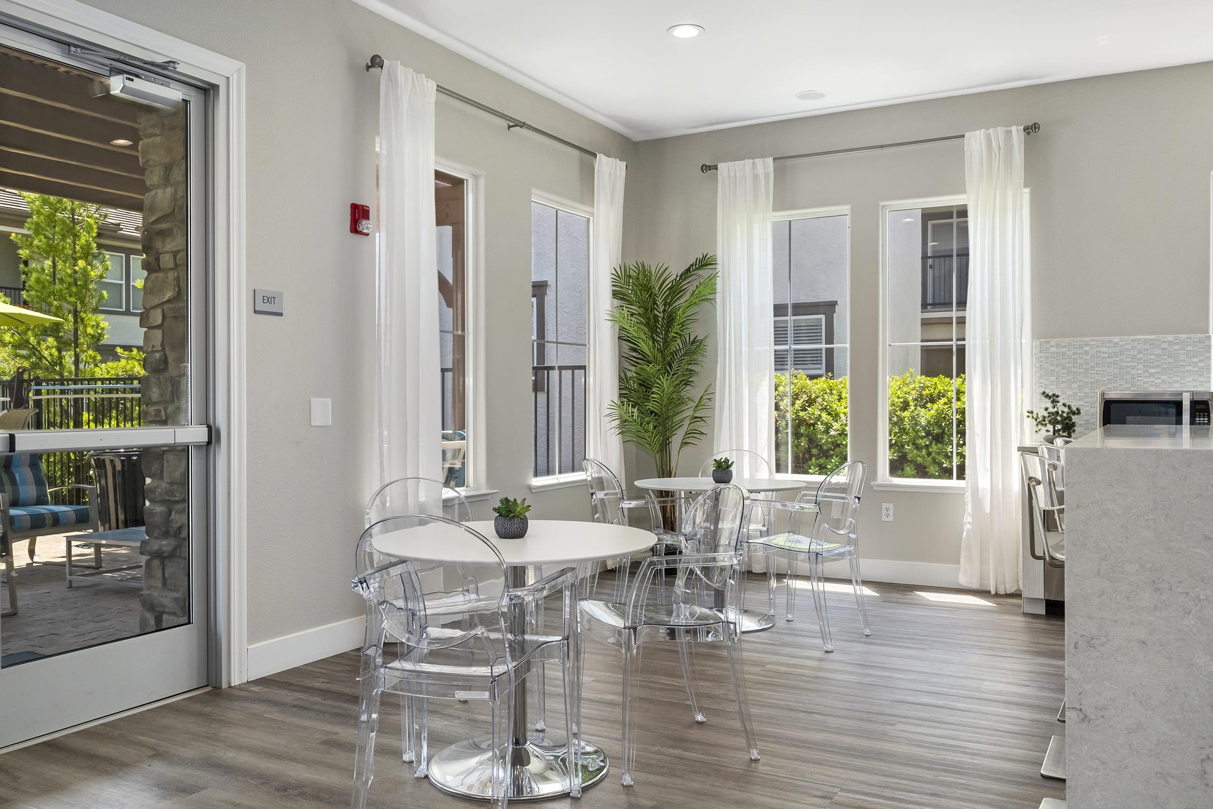 A modern interior of a bright lounge area featuring clear plastic chairs and small round tables. Sunlight streams through large windows adorned with light curtains, and a potted plant adds a touch of greenery. A countertop is visible, enhancing the contemporary atmosphere of the space.