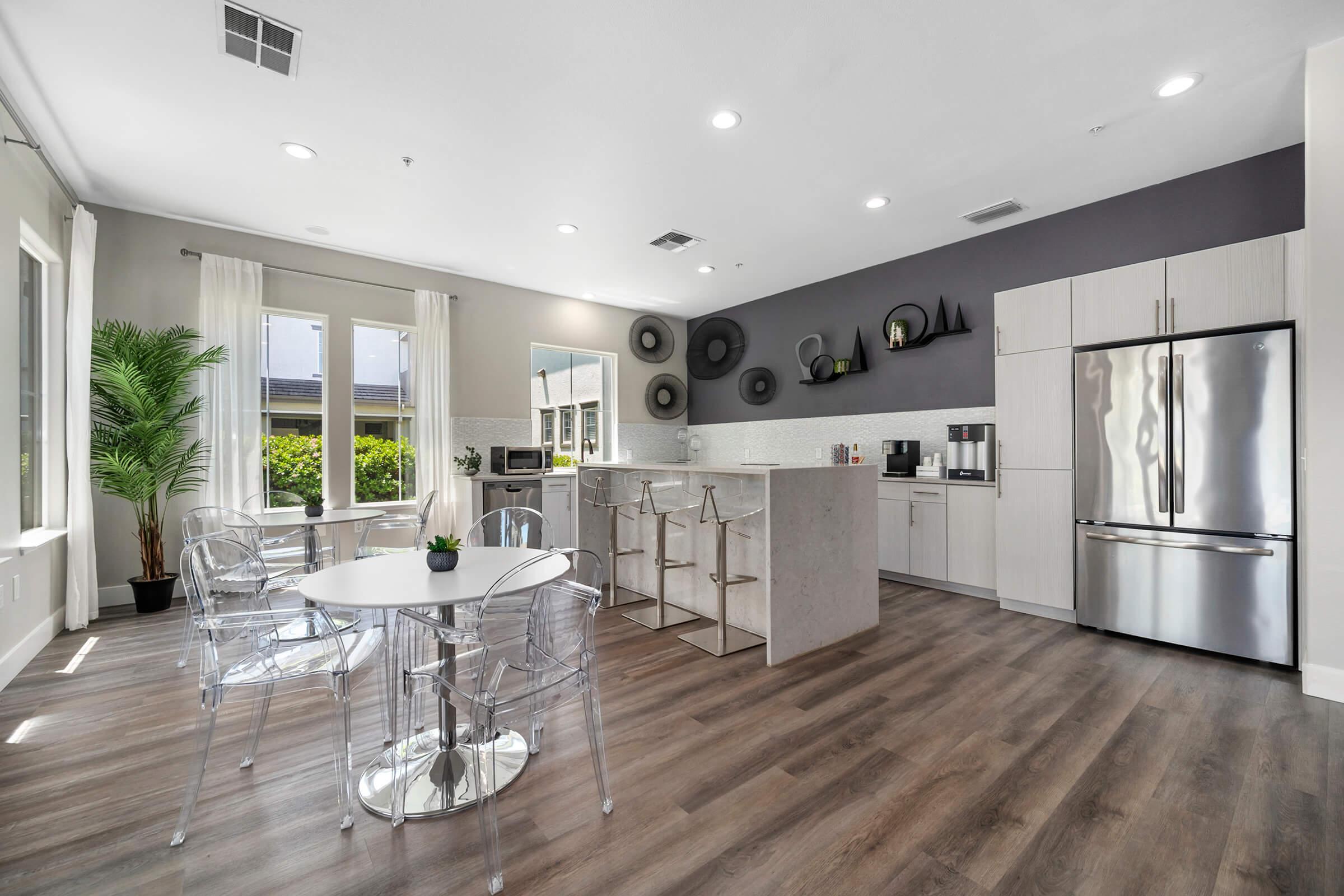 Modern kitchen and dining area featuring a sleek design with white cabinets, stainless steel appliances, and a large island. There are four transparent chairs around a small round table, and a potted plant adds a touch of greenery. Large windows offer natural light and views of the outside.