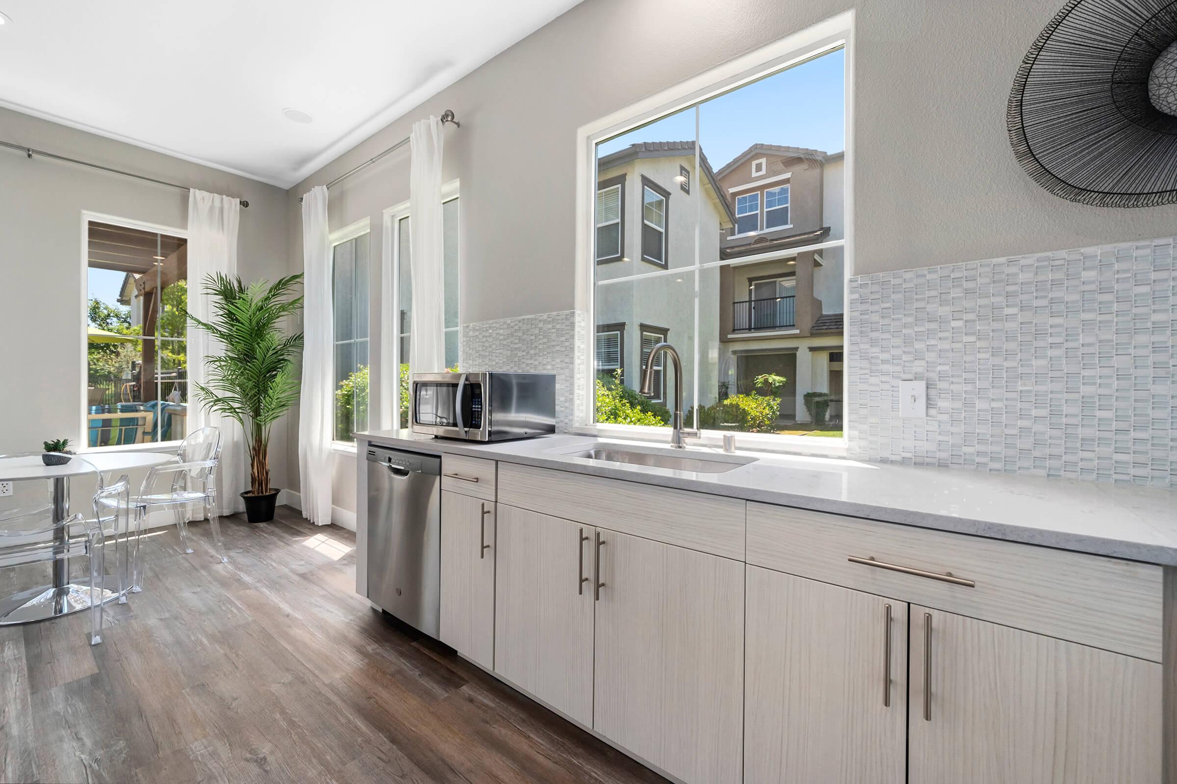 Modern kitchen featuring light wood cabinetry, stainless steel appliances, and a large window that lets in natural light. A dining area with clear chairs and a vibrant green plant is visible, giving a fresh and inviting atmosphere.
