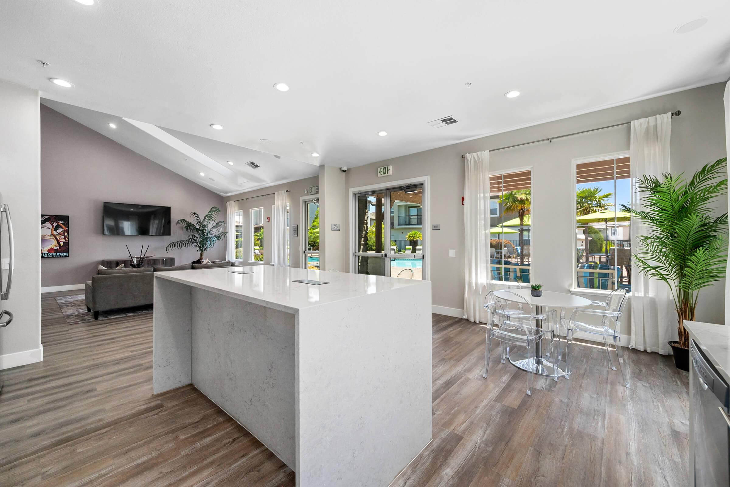 Modern interior of a stylish living space featuring a bright kitchen island with white countertops, a seating area with clear chairs, and a spacious lounge area with a television. Large windows allow natural light, showcasing palm trees outside, contributing to a relaxed and inviting atmosphere.