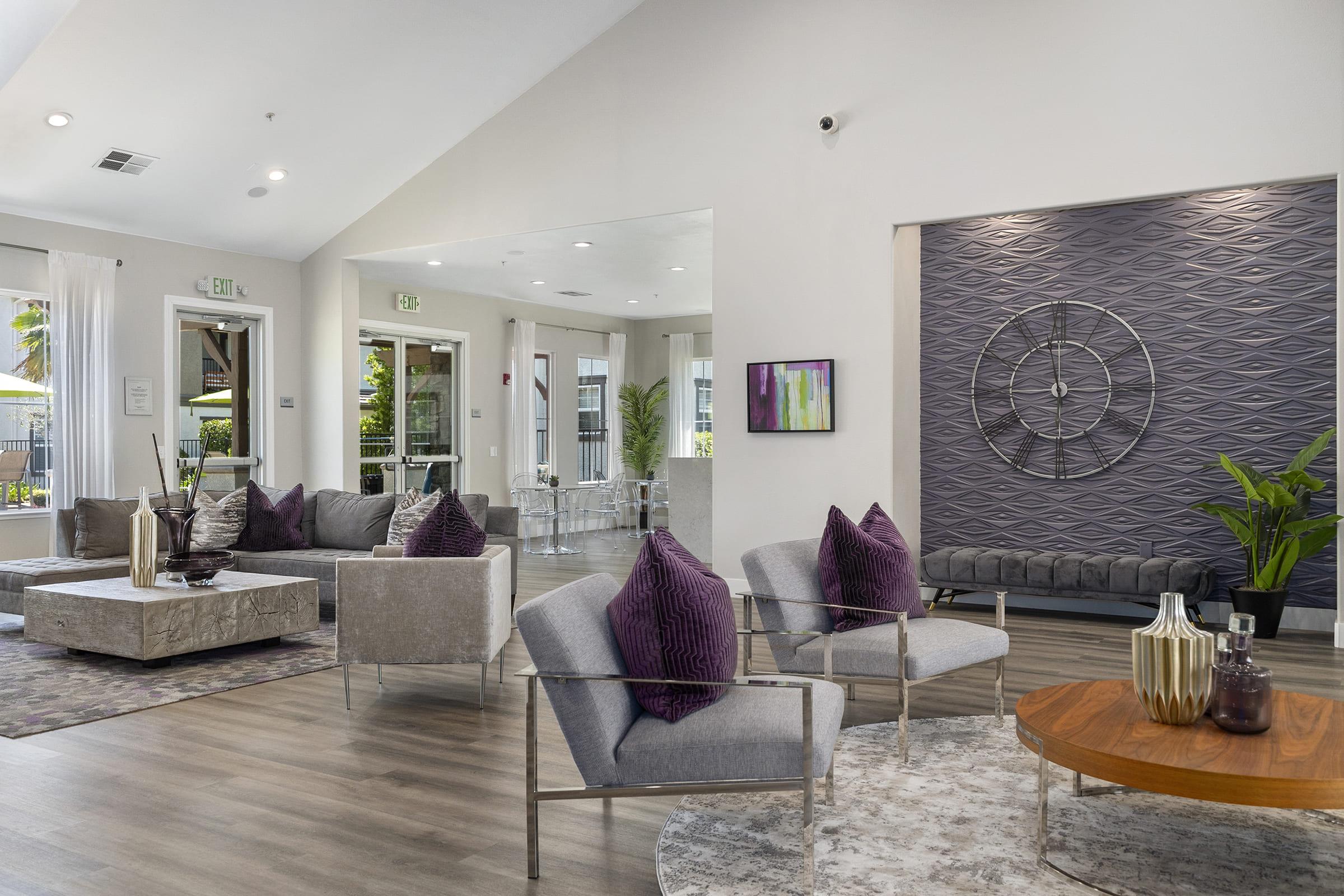 Modern interior of a spacious lounge area featuring stylish furniture. Plush gray chairs with purple accent pillows are arranged around a wooden coffee table. A textured wall with circular decor adds visual interest, while large windows provide natural light and views of greenery outside.