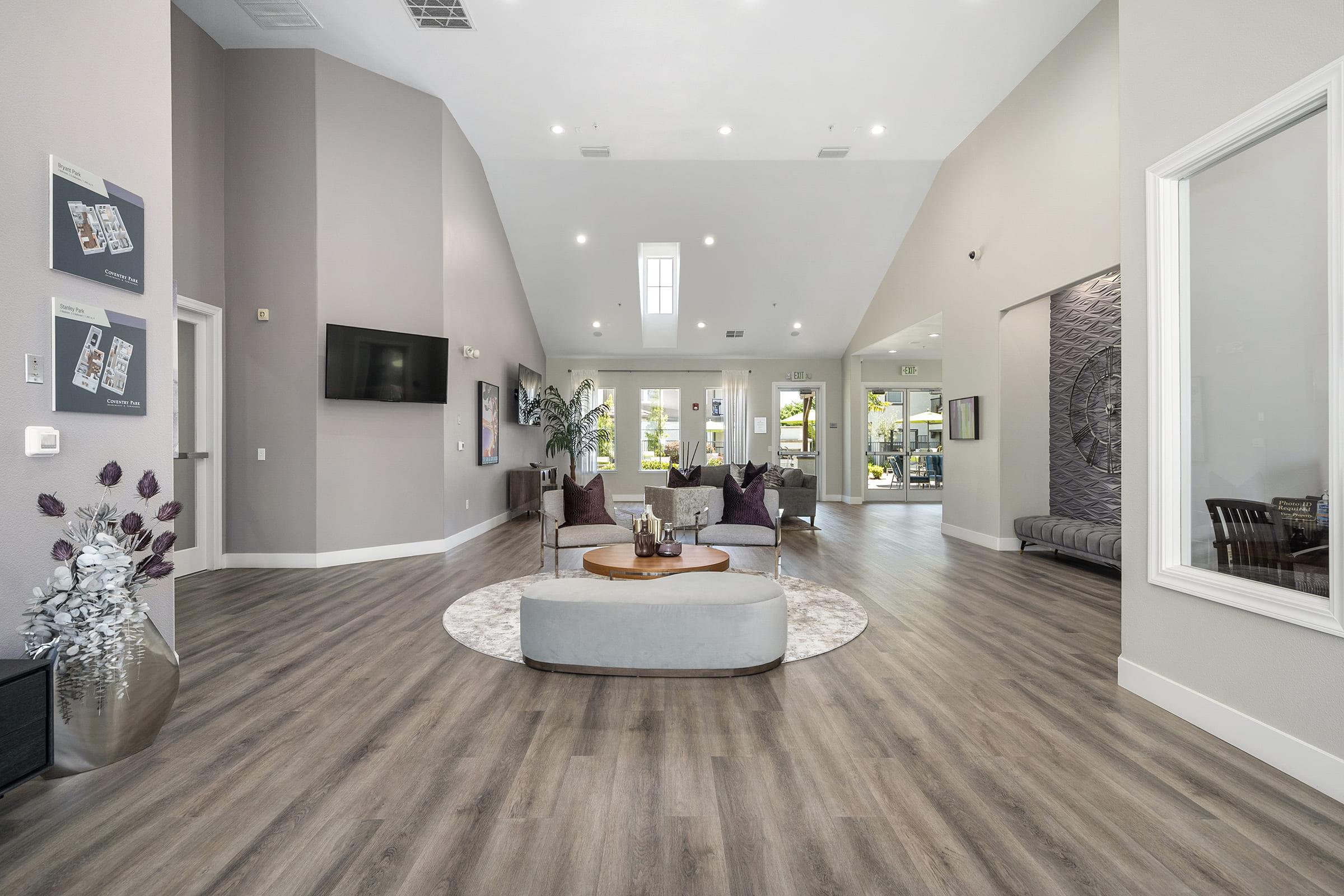 A spacious, modern interior with high ceilings featuring a large, open area. There are comfortable seating arrangements with gray sofas and decorative pillows. A circular area rug sits in the center, and there are decorative plants and wall art. Large windows allow natural light to fill the space.