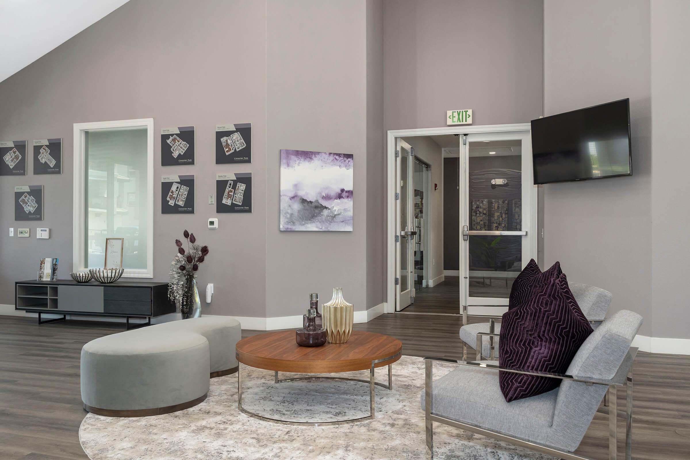 A modern, stylish lounge area featuring gray walls, a circular coffee table with decorative items, and a round ottoman. Two gray chairs with purple cushions are arranged around the table. The space includes framed artwork on the walls, a TV, and glass doors leading to an exit. A warm, inviting atmosphere is created by the decor.
