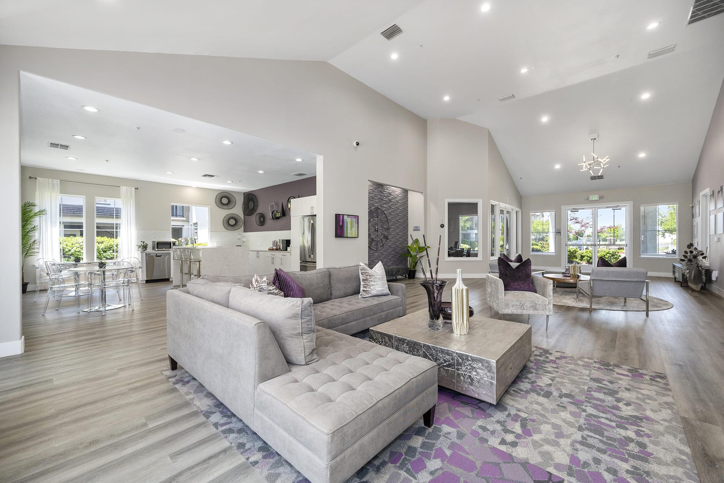 A spacious living area featuring a modern gray sectional sofa with decorative pillows, a coffee table, and a colorful area rug. Large windows allow natural light, showcasing a view of greenery outside. The kitchen is visible in the background, along with dining chairs and a minimalist decor style.