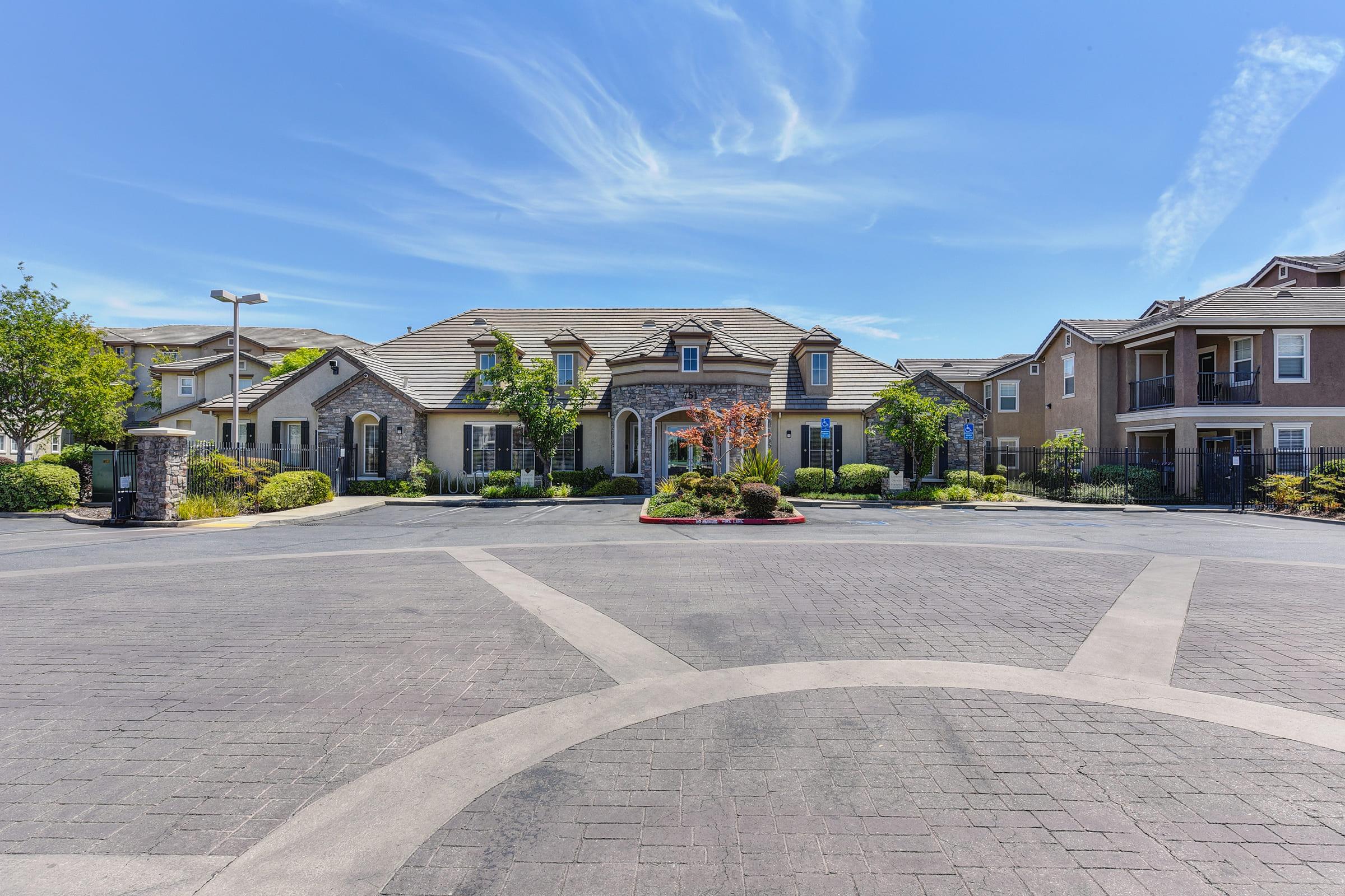 A landscaped entrance to an apartment complex featuring two buildings with a central circular driveway. The scene is illuminated by bright blue skies and includes green trees, shrubs, and decorative plants along the pathway.