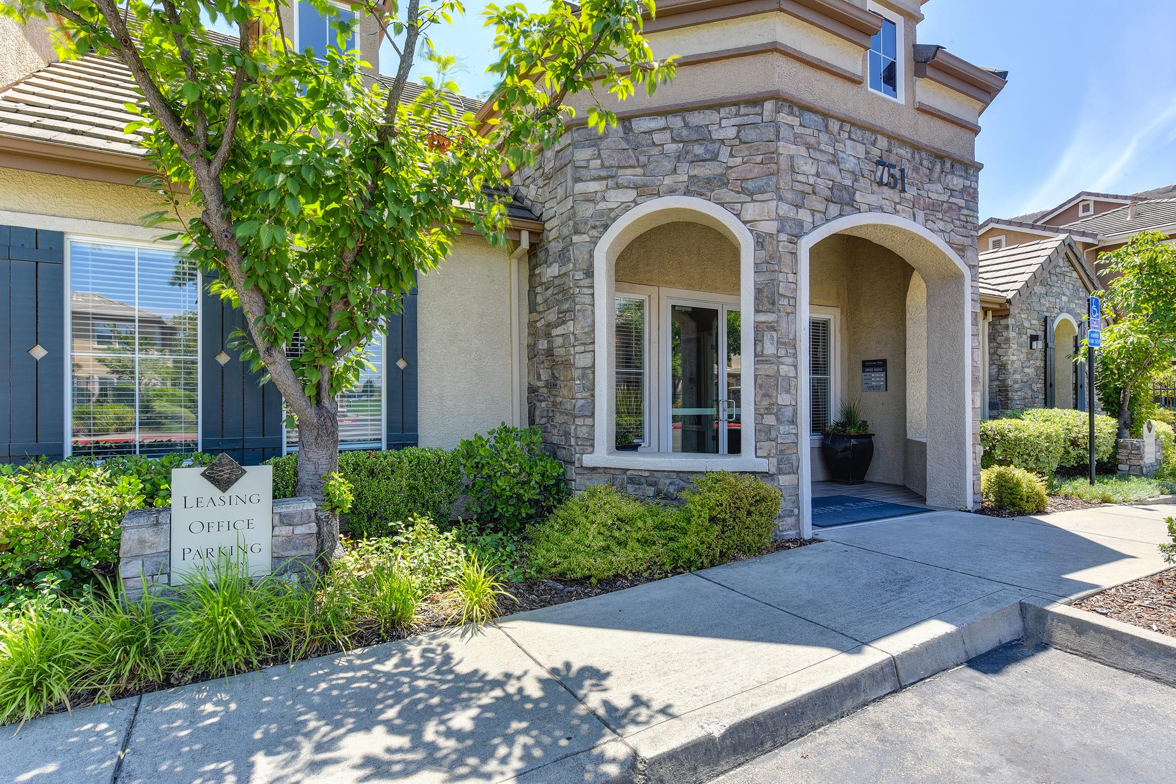 Exterior view of a leasing office featuring stone archways, large windows, and well-maintained landscaping. A sign indicating "Leasing Office Parking" is visible near the entrance, and the bright, sunny atmosphere highlights the modern architecture and greenery surrounding the building.