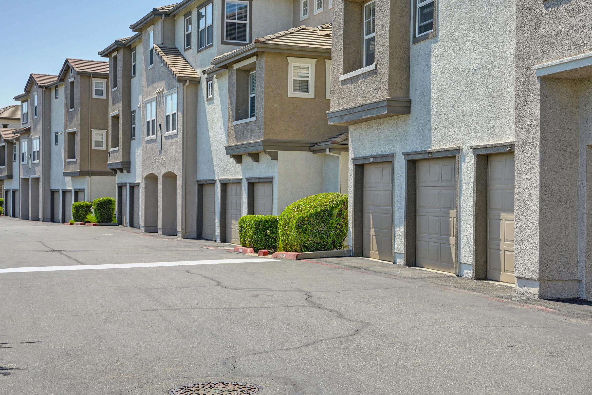 A row of multi-story residential buildings with beige and gray exteriors. The structures have garages with closed doors and manicured shrubs in front. The pavement is smooth and there are marked parking areas. A clear blue sky is visible overhead, indicating a bright day.