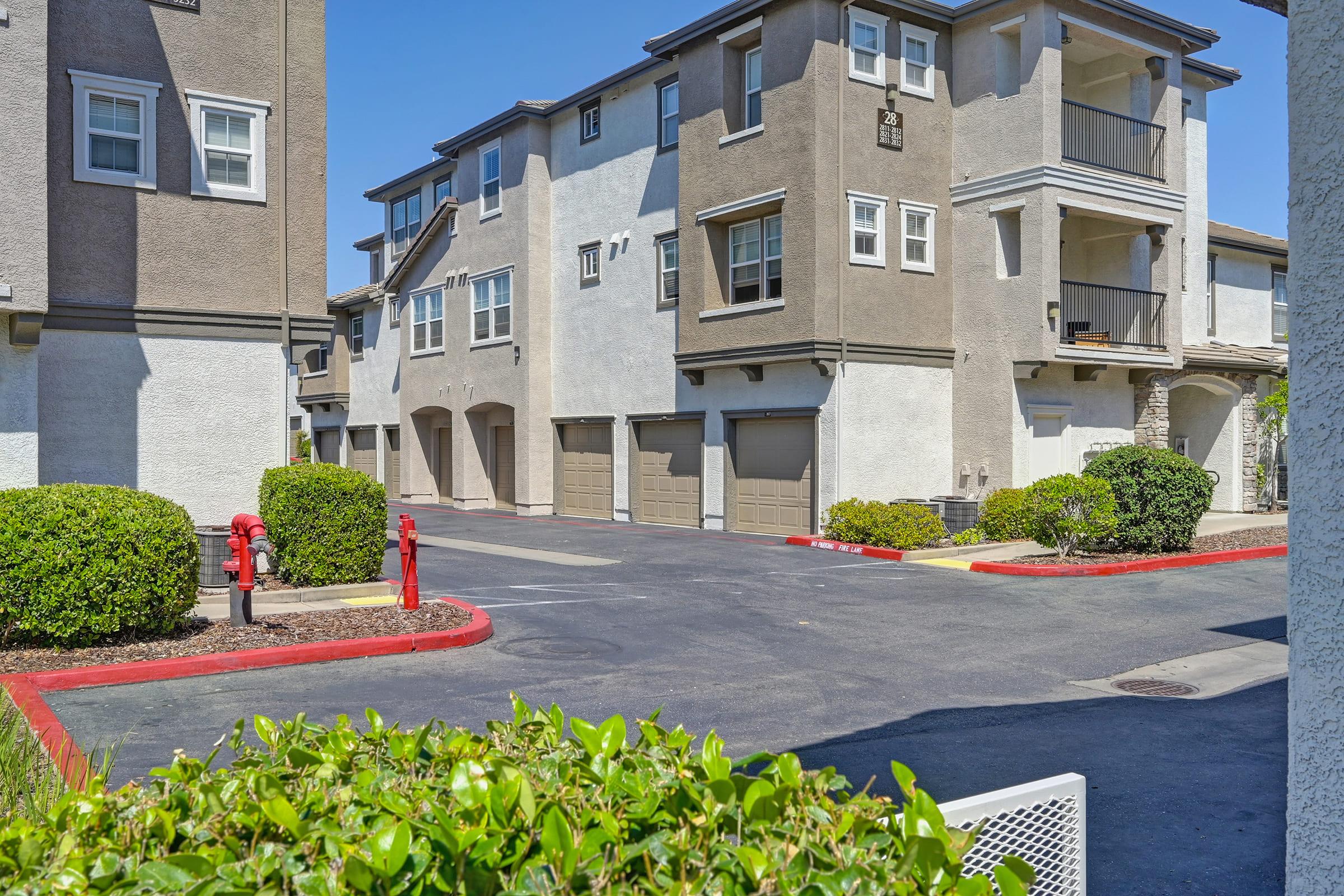 A view of a residential area featuring multiple two- and three-story apartment buildings. The scene includes garages, landscaped hedges, and a parking area marked with red curbs. There are fire hydrants visible, and the sky is clear and blue.