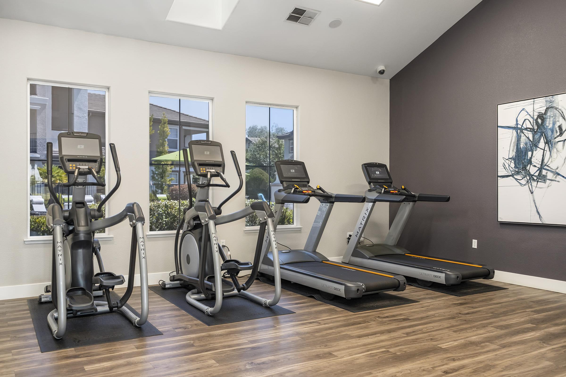A modern gym interior featuring three exercise machines: two treadmills and one elliptical trainer. The room has large windows allowing natural light, with a view of greenery outside. The walls are painted in neutral tones, and there's abstract artwork on one wall. The flooring is wooden, adding warmth to the space.