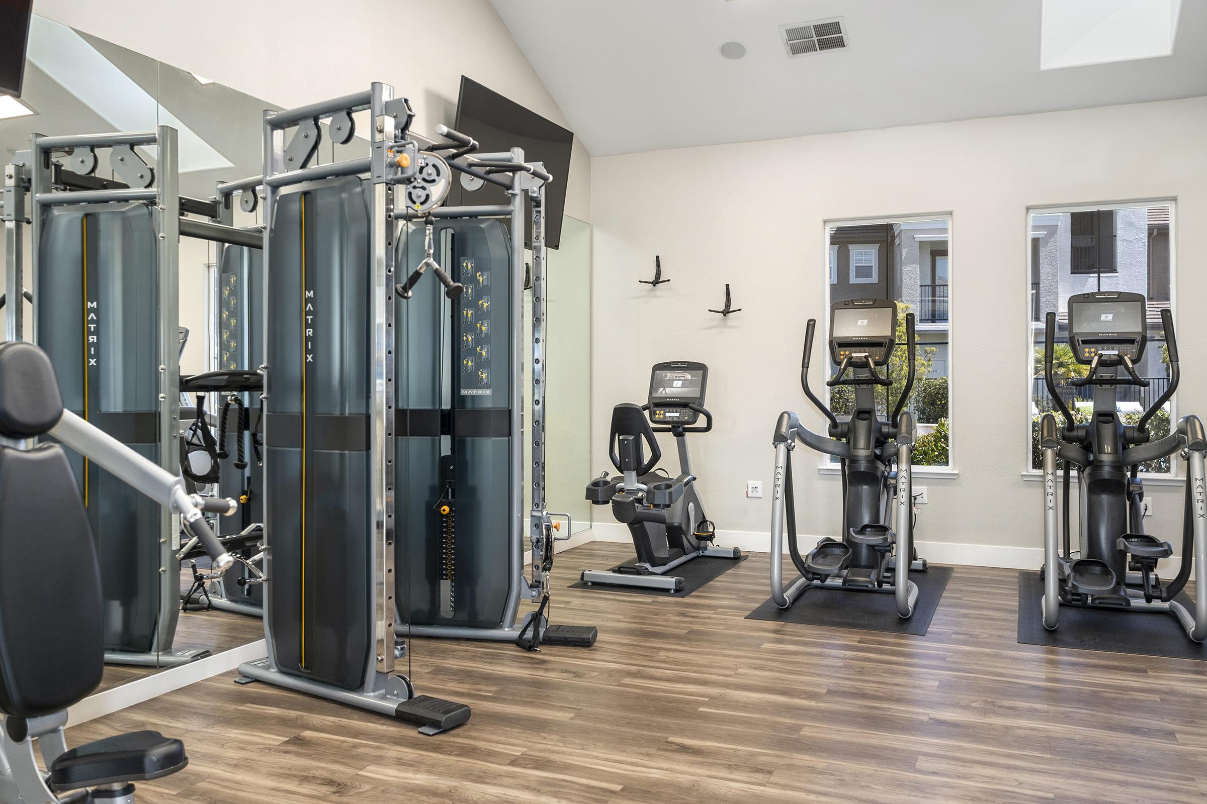 A well-lit gym featuring various exercise equipment including a cable machine, stationary bike, and elliptical trainer. Large windows provide natural light, and the floor is made of wooden planks. The space is clean and organized, encouraging fitness activities.