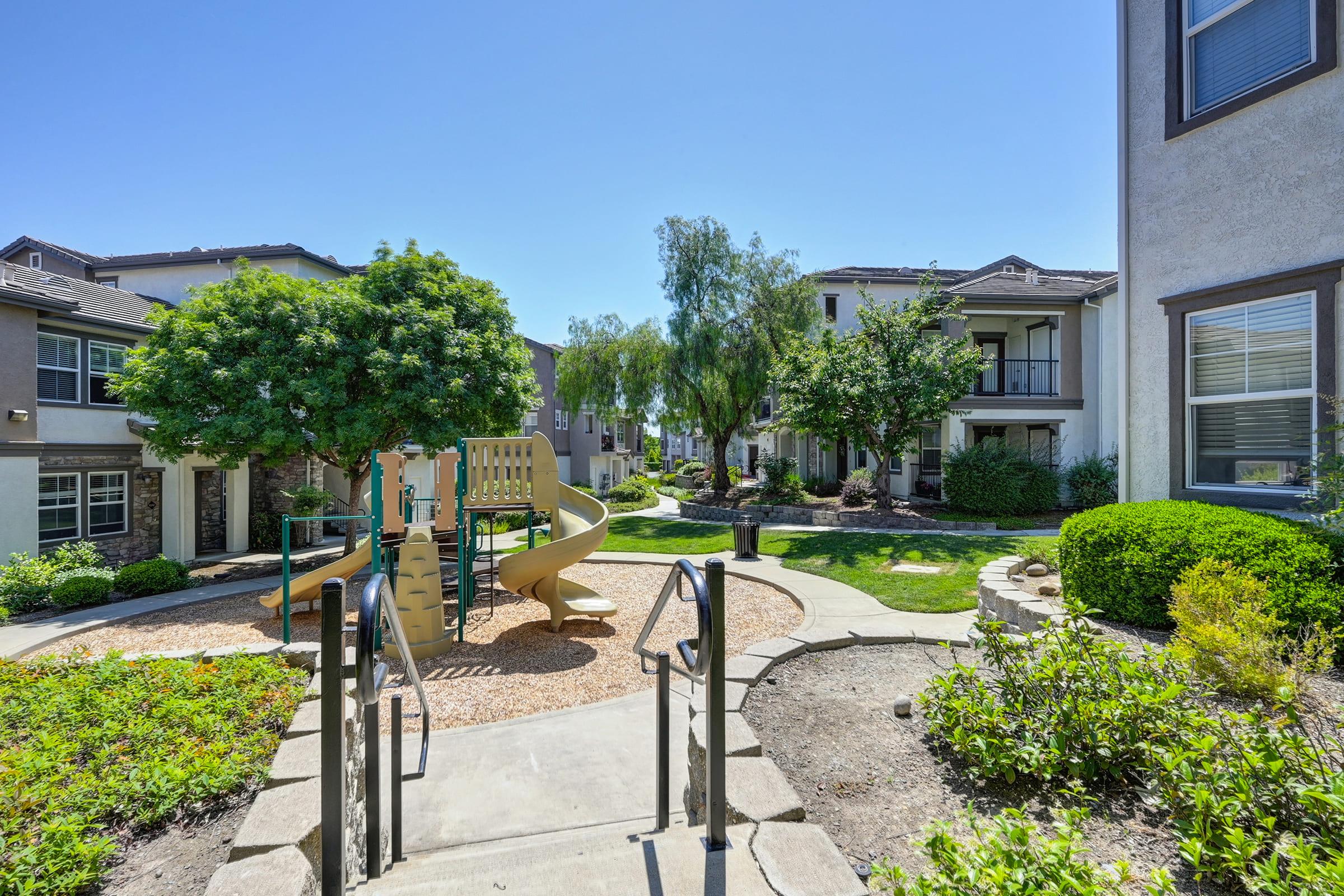 View of a landscaped courtyard in a residential area featuring a playground with a slide, surrounded by green trees and manicured bushes. The area is sunny and well-maintained, with pathways leading to several apartment buildings