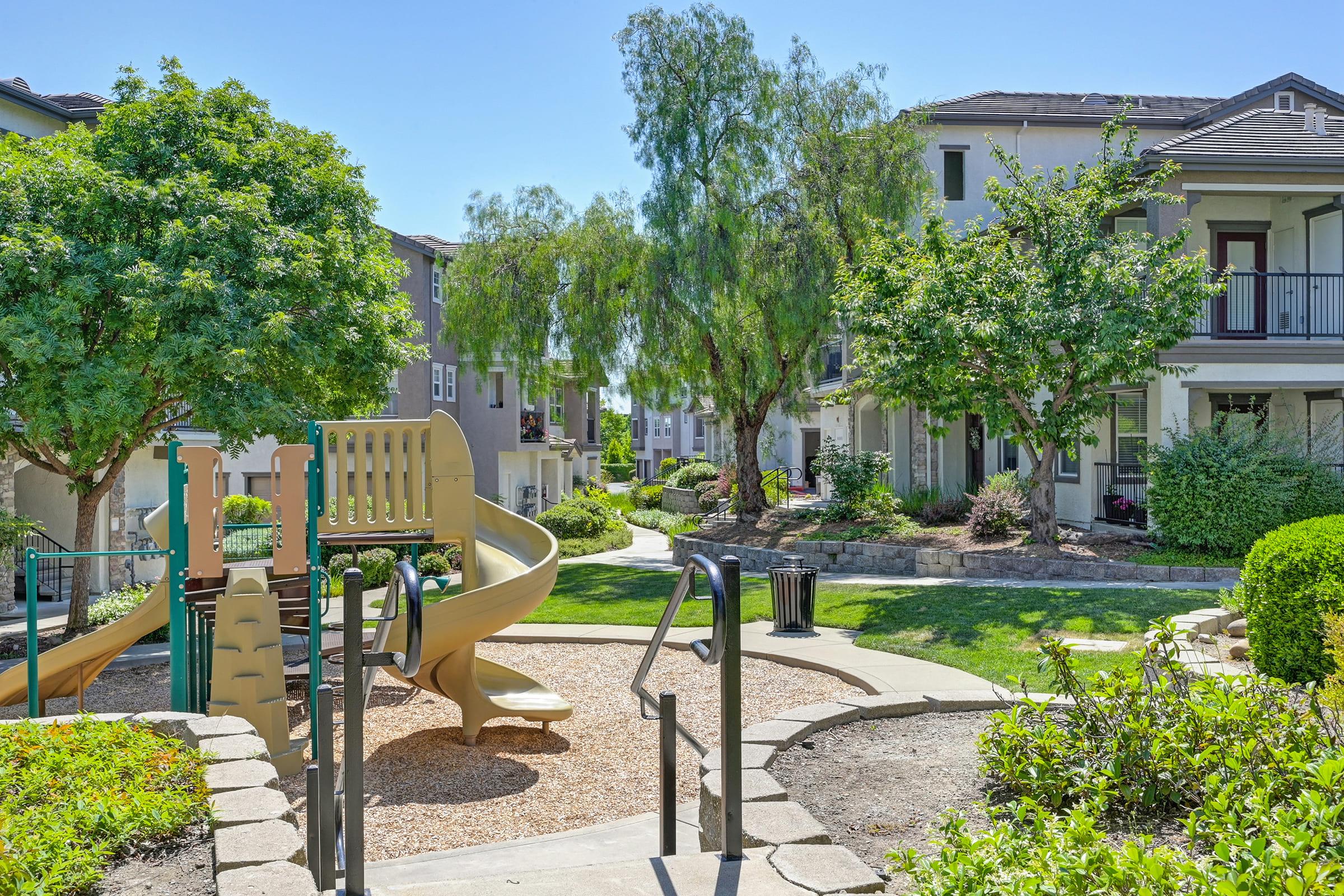 A playground with a slide surrounded by green trees and shrubs, set in a residential area. The scene features pathways, benches, and well-maintained landscaping, providing a welcoming outdoor space for families and children. Clear blue sky overhead enhances the cheerful atmosphere.