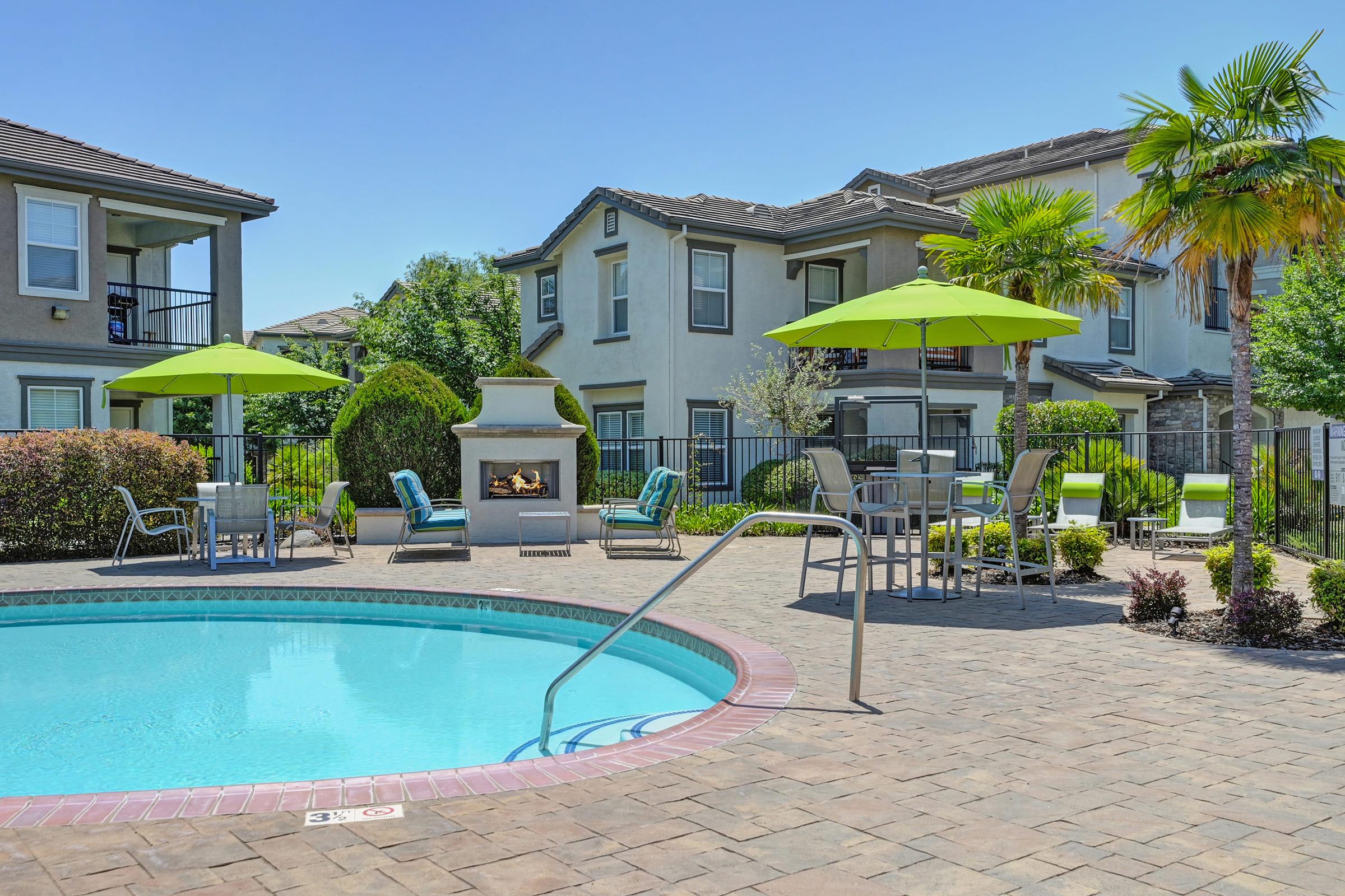 A sunny poolside area featuring a clear blue swimming pool, surrounded by lounge chairs and green umbrellas. In the background, there are modern apartment buildings and lush landscaping, including palm trees and shrubs. A cozy outdoor fireplace adds to the inviting atmosphere.