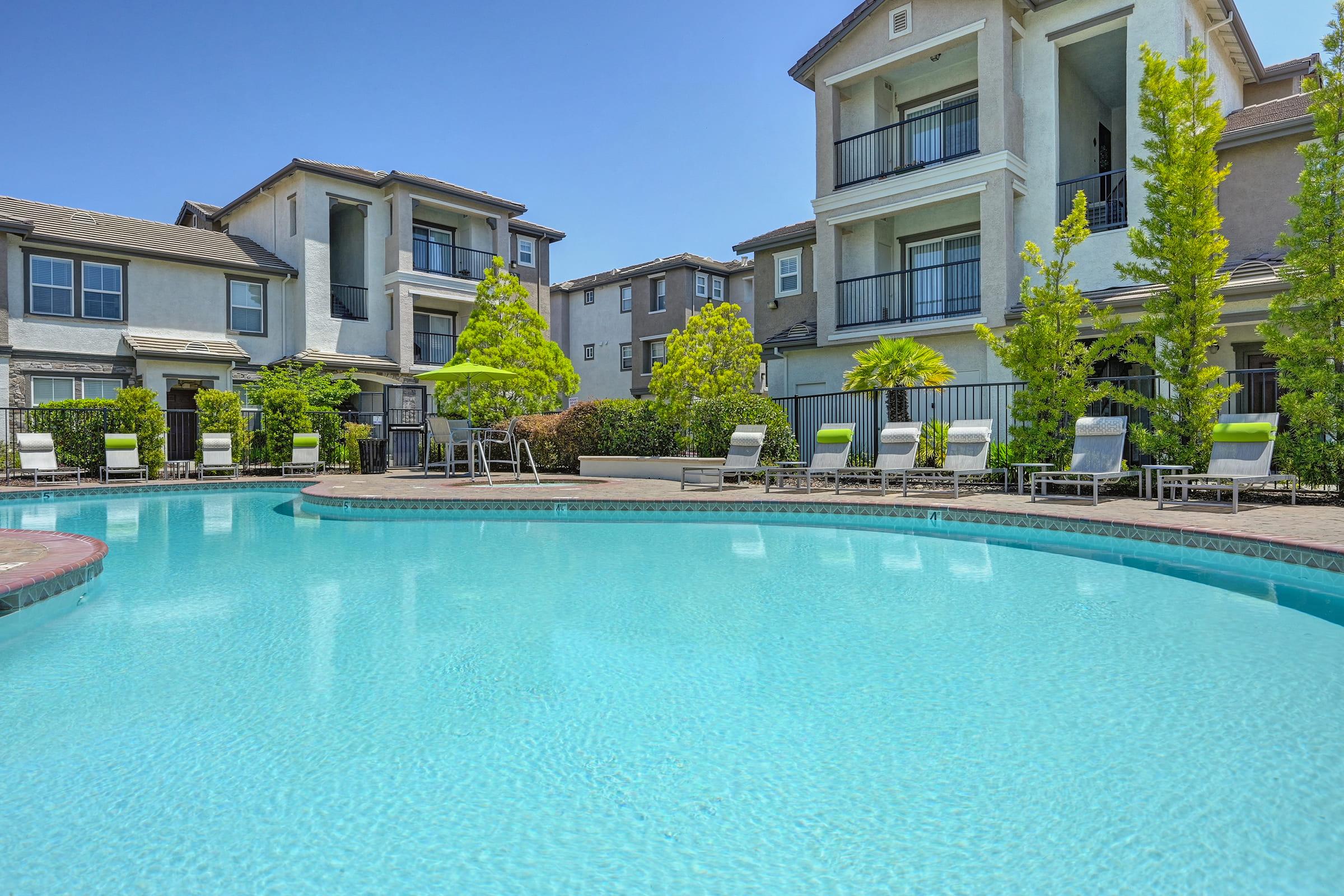 A clear, blue swimming pool surrounded by lounge chairs and green trees, with modern apartment buildings in the background. The sky is bright and sunny, creating a relaxing and inviting atmosphere.