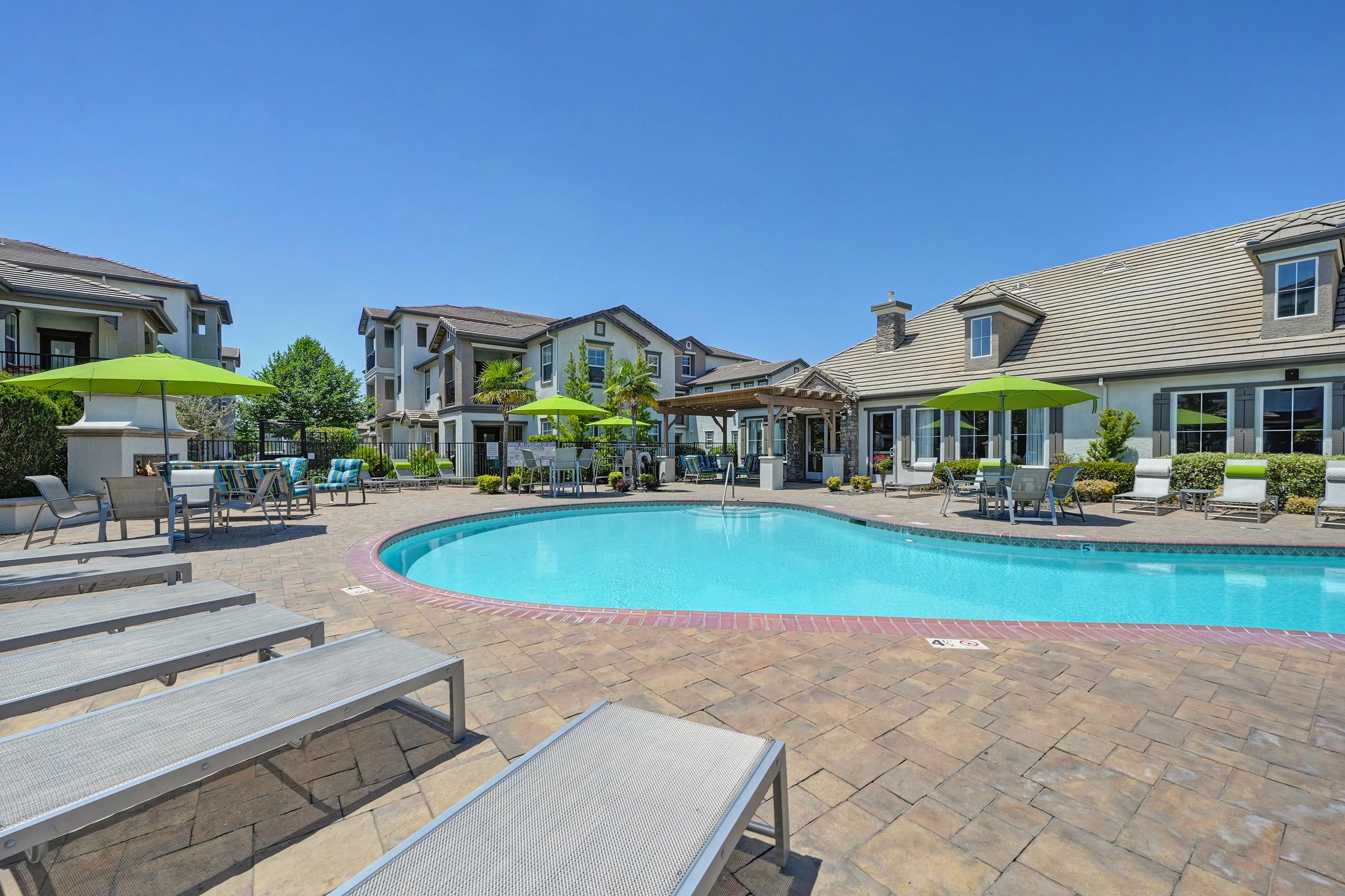 A sunny outdoor swimming pool area with lounge chairs and umbrellas. Surrounding the pool are well-maintained pathways and landscaping, with residential buildings in the background. The clear blue sky adds to the inviting atmosphere of the space.