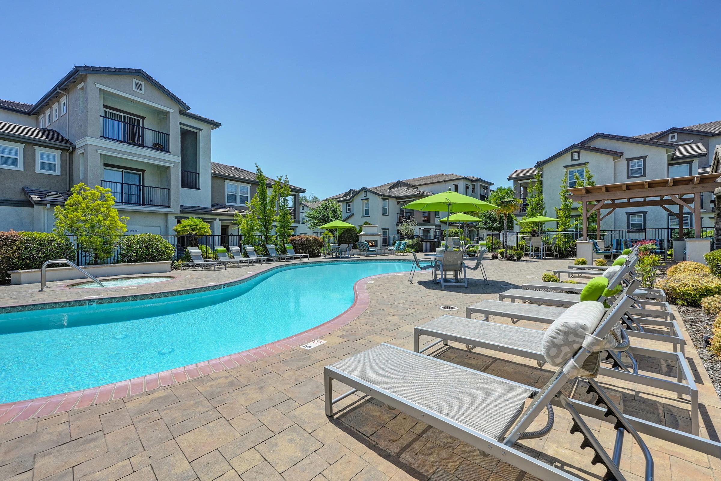 A vibrant outdoor pool area with a sparkling blue pool surrounded by lounge chairs. Green umbrellas provide shade, and well-maintained landscaping adds to the inviting atmosphere. Several modern apartment buildings are visible in the background under a clear blue sky.