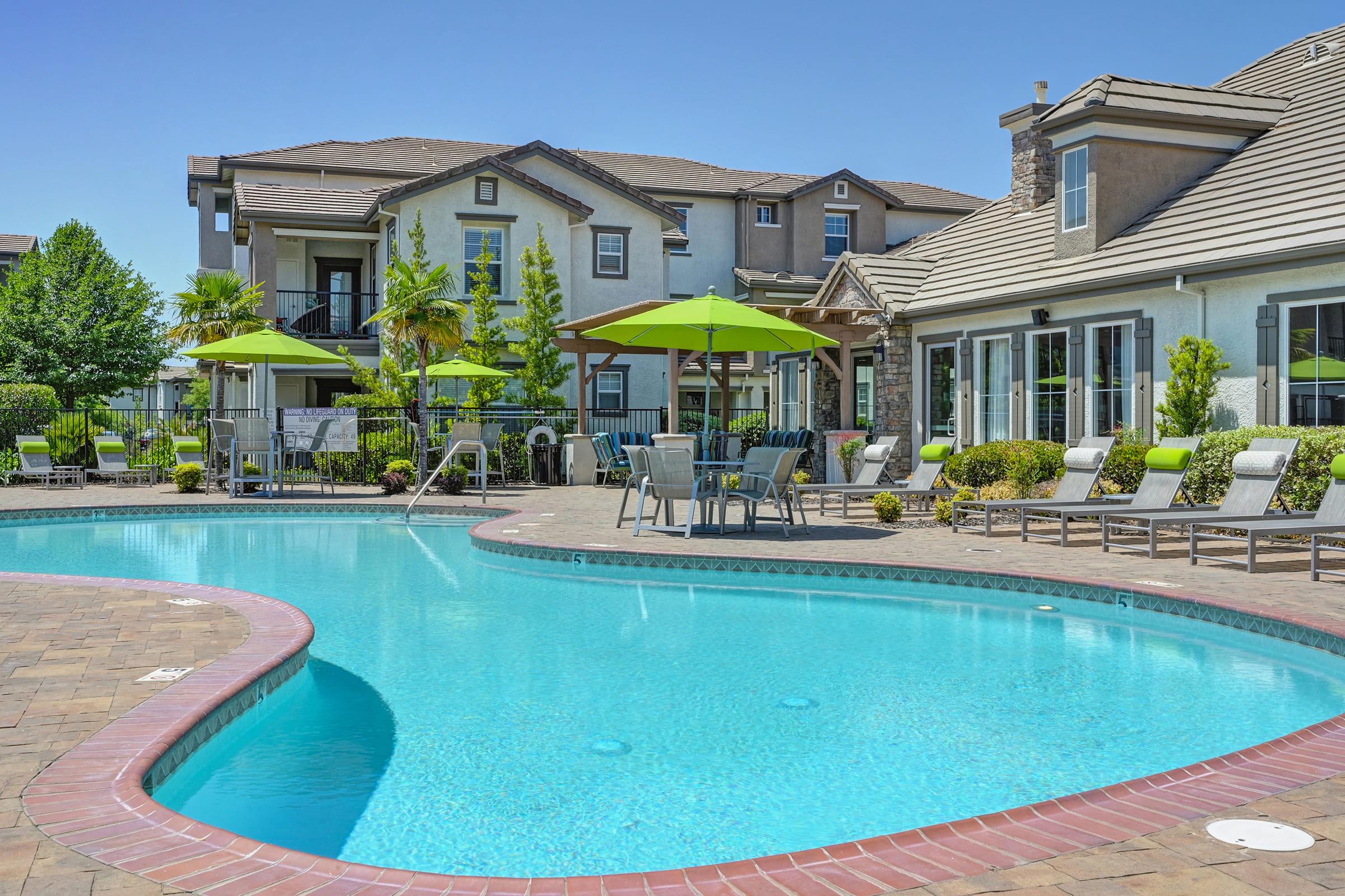 A sparkling blue swimming pool surrounded by lounge chairs and green umbrellas, with a well-maintained apartment complex in the background. Lush plants and trees enhance the inviting outdoor space, under a clear blue sky. Perfect for relaxation and leisure.